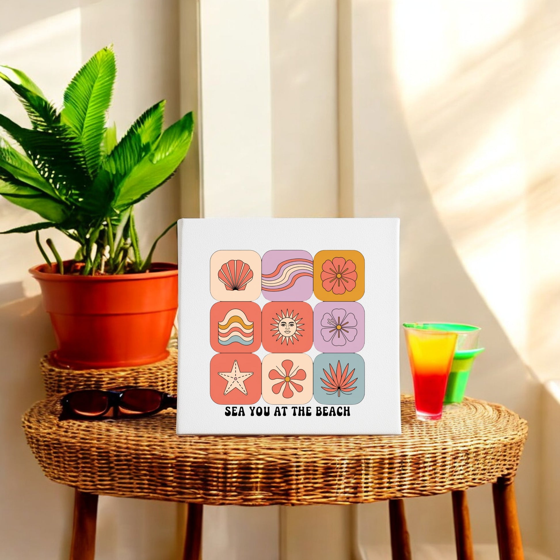 A rattan table holds sunglasses, a tropical drink, and a plant beside Mirabilia Boutique's Sea You at the Beach Canvas – Retro Coastal Wall Art. Sunlight enhances the relaxed retro summer vibe.