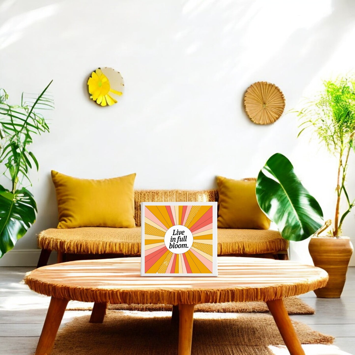 A bright, airy living room with wicker furniture, yellow cushions, green plants, and a round wicker coffee table. Sunlight highlights the Mirabilia Boutique “Live in Full Bloom” Summer Sunburst Canvas displayed on the table.