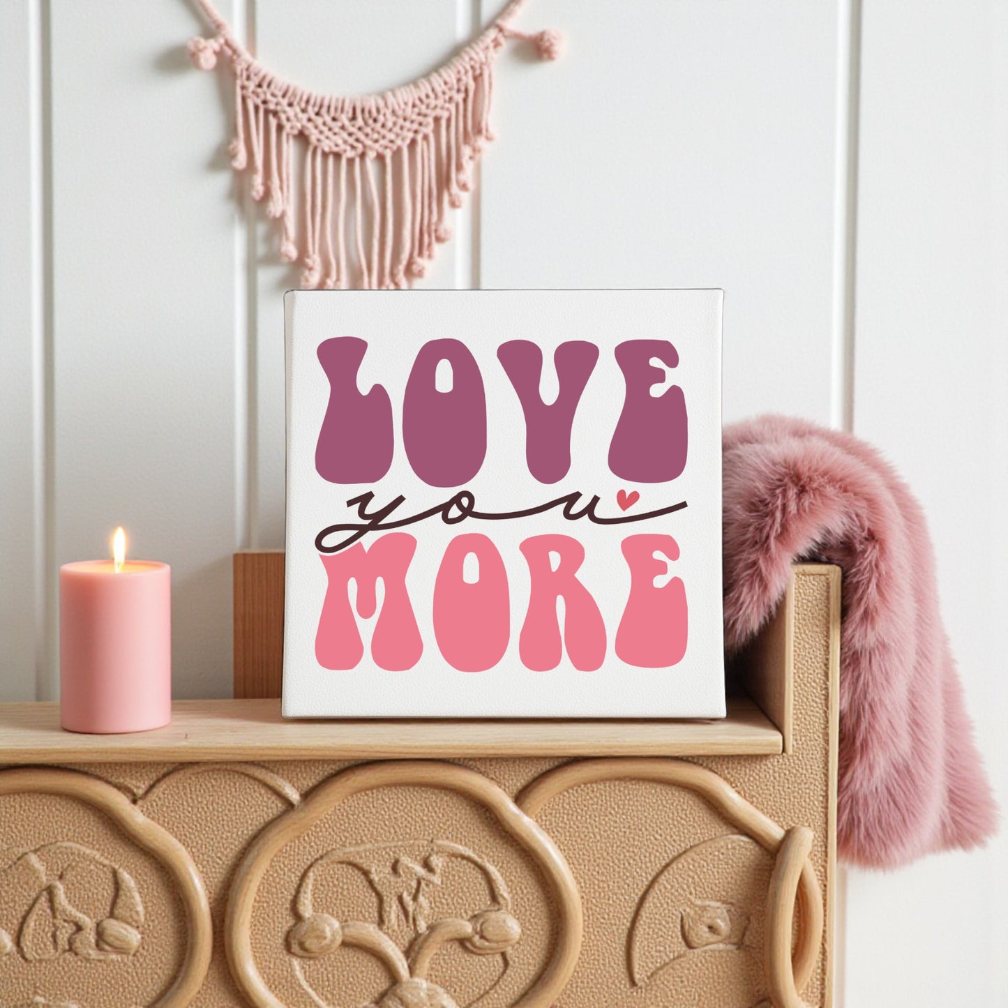 The Love You More 8x8 Canvas by Mirabilia Boutique features bold purple and pink retro letters on white, displayed on a carved wooden surface with a pink candle—groovy wall art perfect for retro decor lovers, with a macrame hanging in the background.