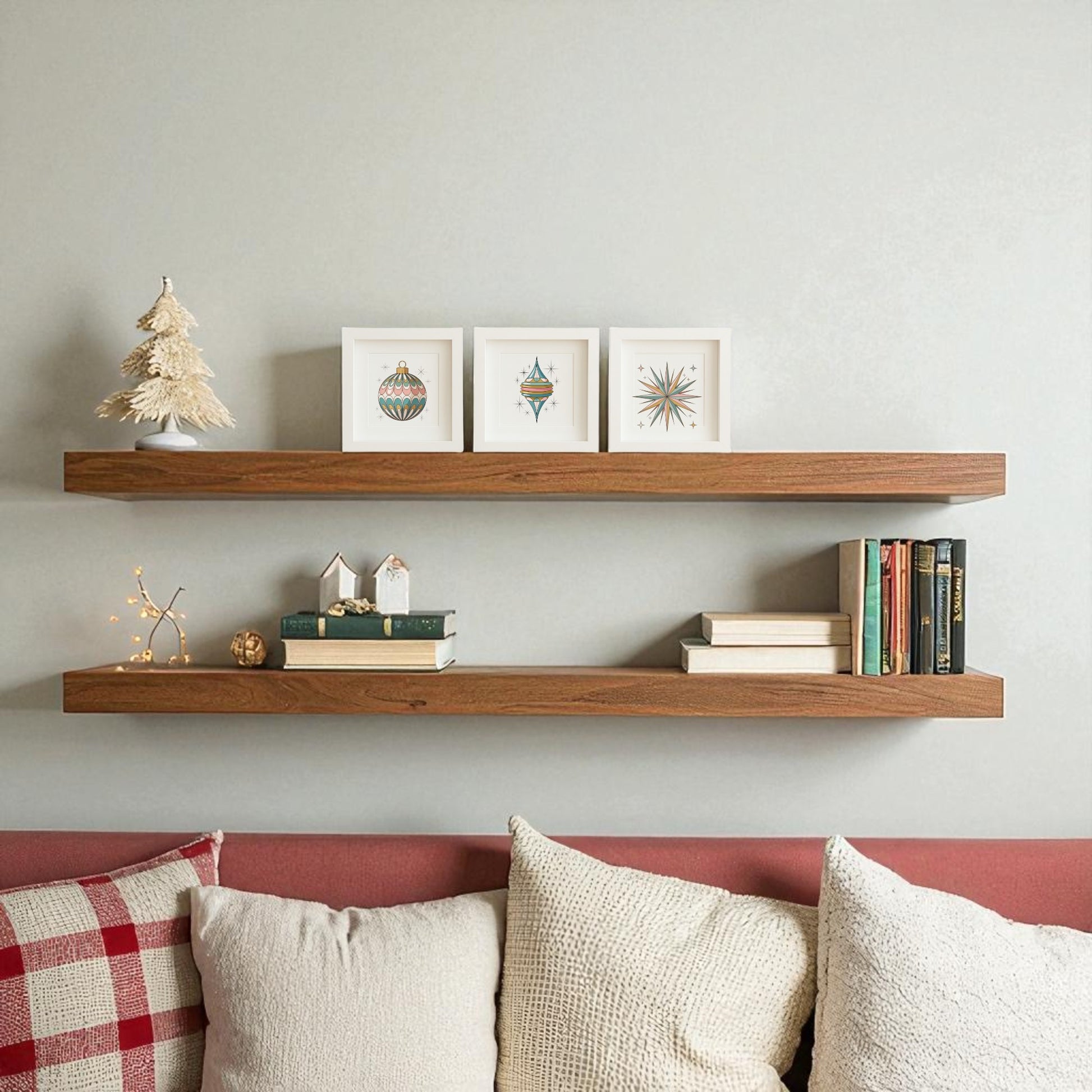 Two wooden floating shelves on a light wall showcase small framed art, books, a miniature tree, and the Retro Ornament Trio Mid-Century Holiday Wall Art Set by Mirabilia Boutique above a sofa with white and red cushions.