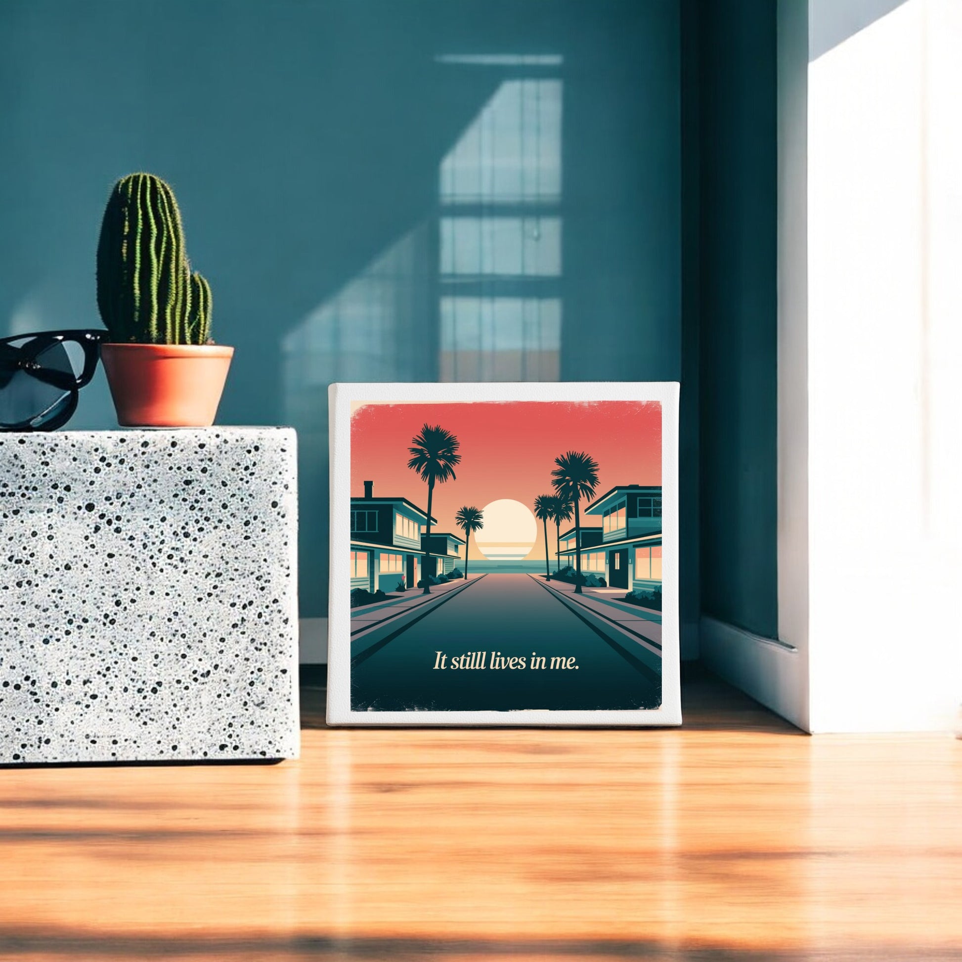 The Mirabilia Boutique "It Still Lives in Me Canvas – Retro Sunset Memory Art" features palm trees, buildings, and a sunset with the words “It still lives in me,” displayed on a wooden floor by terrazzo, cactus, and sunglasses.