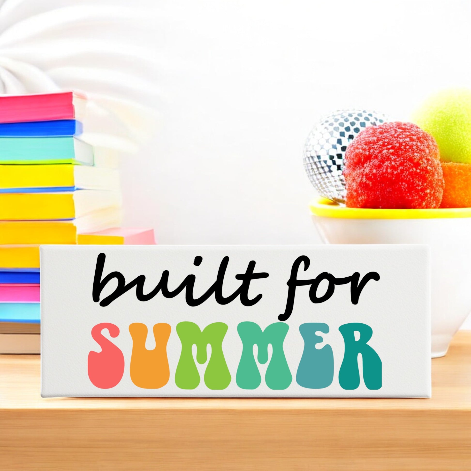 The Built for Summer Canvas – Retro Shelf Sign by Mirabilia Boutique features playful, colorful letters on white—perfect for summer decor. Displayed with stacked rainbow books and a bowl of bright candies, it brightens any shelf.