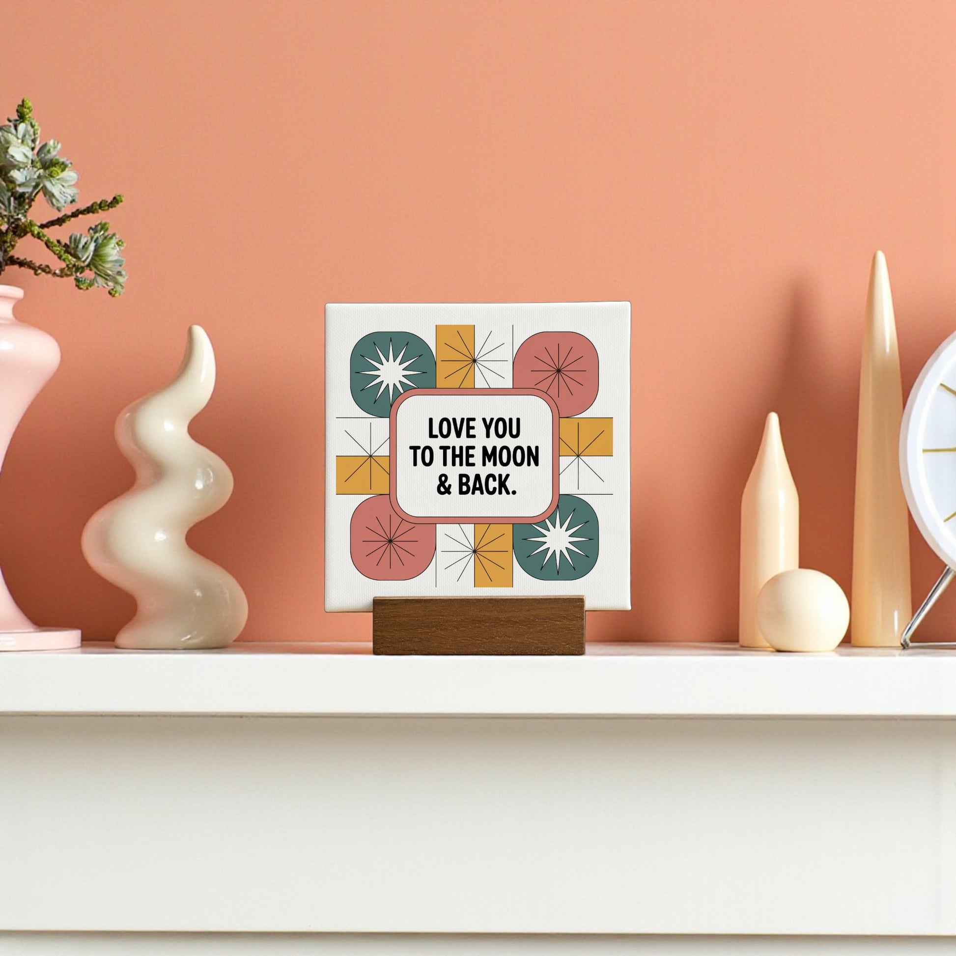 Add a retro vibe to your mantel with the Mirabilia Boutique "Love You to the Moon & Back" 4x4 mini canvas sign, featuring colorful starbursts and displayed alongside vases, a clock, and a small plant on a peach wall.