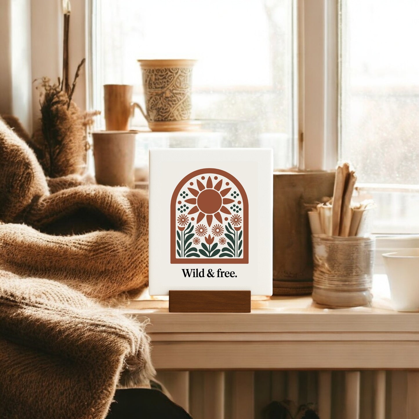 A cozy nook with a brown knitted blanket, cups, and Mirabilia Boutique’s "Wild & Free Mini Sign – Sunflower Boho Desk Art" rests on a windowsill, sunlight streaming in to create a warm, nature-inspired boho vibe.