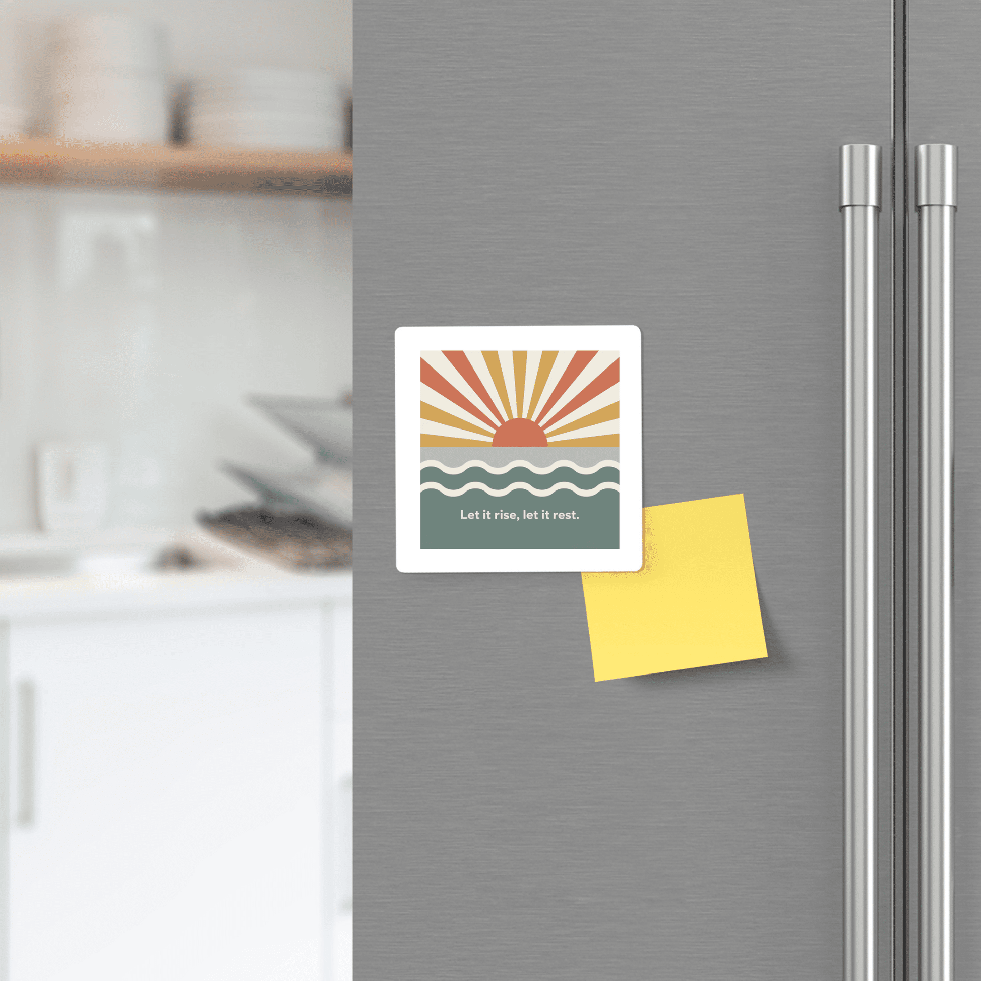 A refrigerator door displays the Let It Rise, Let It Rest Magnet – Sunrise Affirmation by Mirabilia Boutique, featuring a stylized sunrise and waves above a blank yellow sticky note. A kitchen scene is visible in the background.