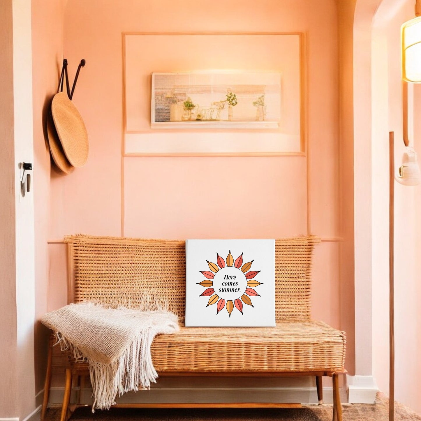 A cozy entryway with peach walls features a wicker bench with a cream throw and Mirabilia Boutique's "Here Comes Summer Canvas – Retro Sunburst Wall Art." Above, a hat hangs on the wall.