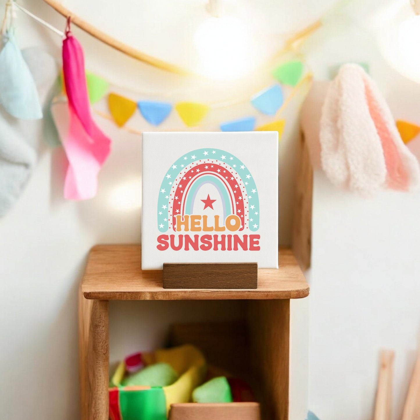 The Mirabilia Boutique Hello Sunshine Mini Canvas Sign with Stand features a pastel rainbow and star design, displayed on a small shelf. Colorful fabric and triangular flag banners decorate the softly lit background.