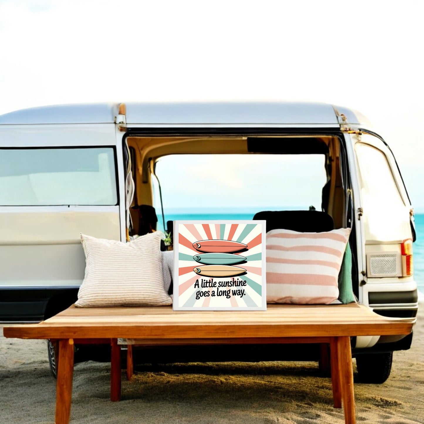A van parked on a beach with its side door open. In front, a wooden bench holds two pillows and Mirabilia Boutique’s "A Little Sunshine Canvas – Retro Surf Summer Vibes" with ocean views in the background.