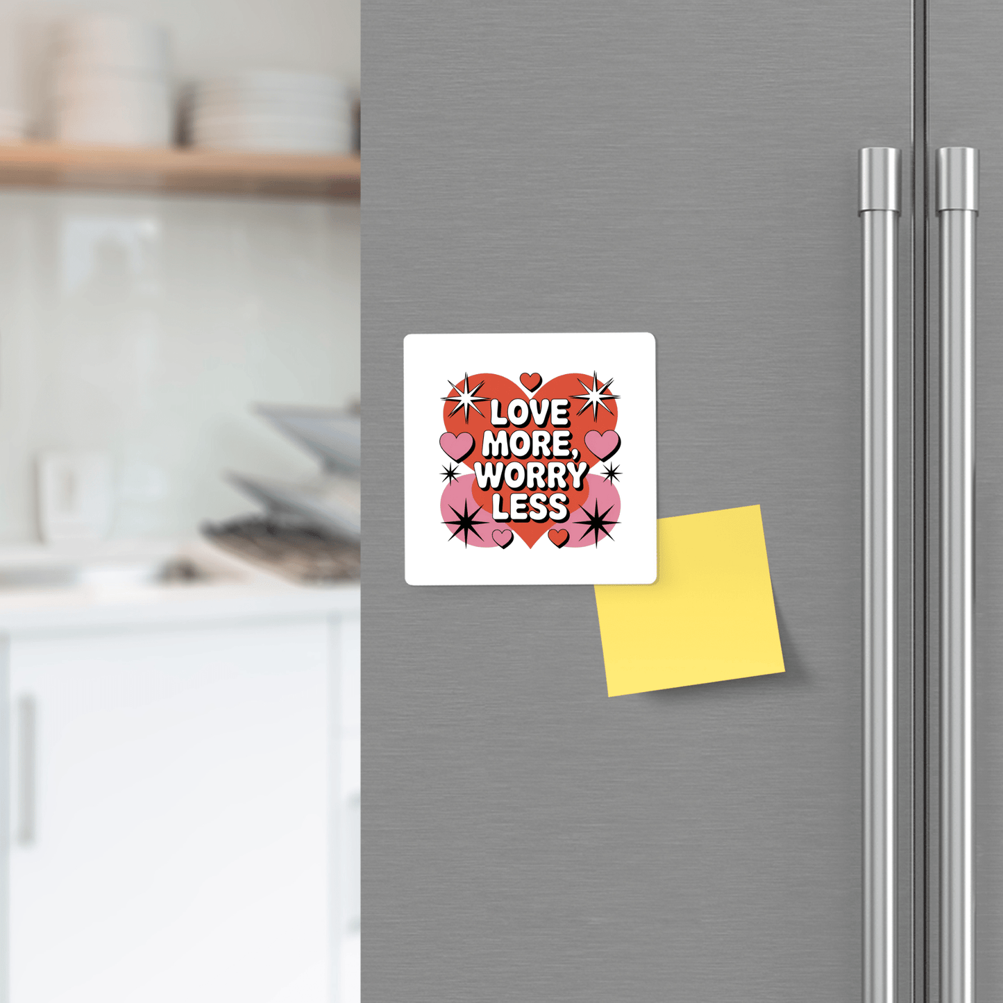 The Love More Worry Less Magnet by Mirabilia Boutique adds retro Valentine charm to your fridge, with bold lettering, hearts, and stars. A yellow sticky note sits below, kitchen shelves in the background.