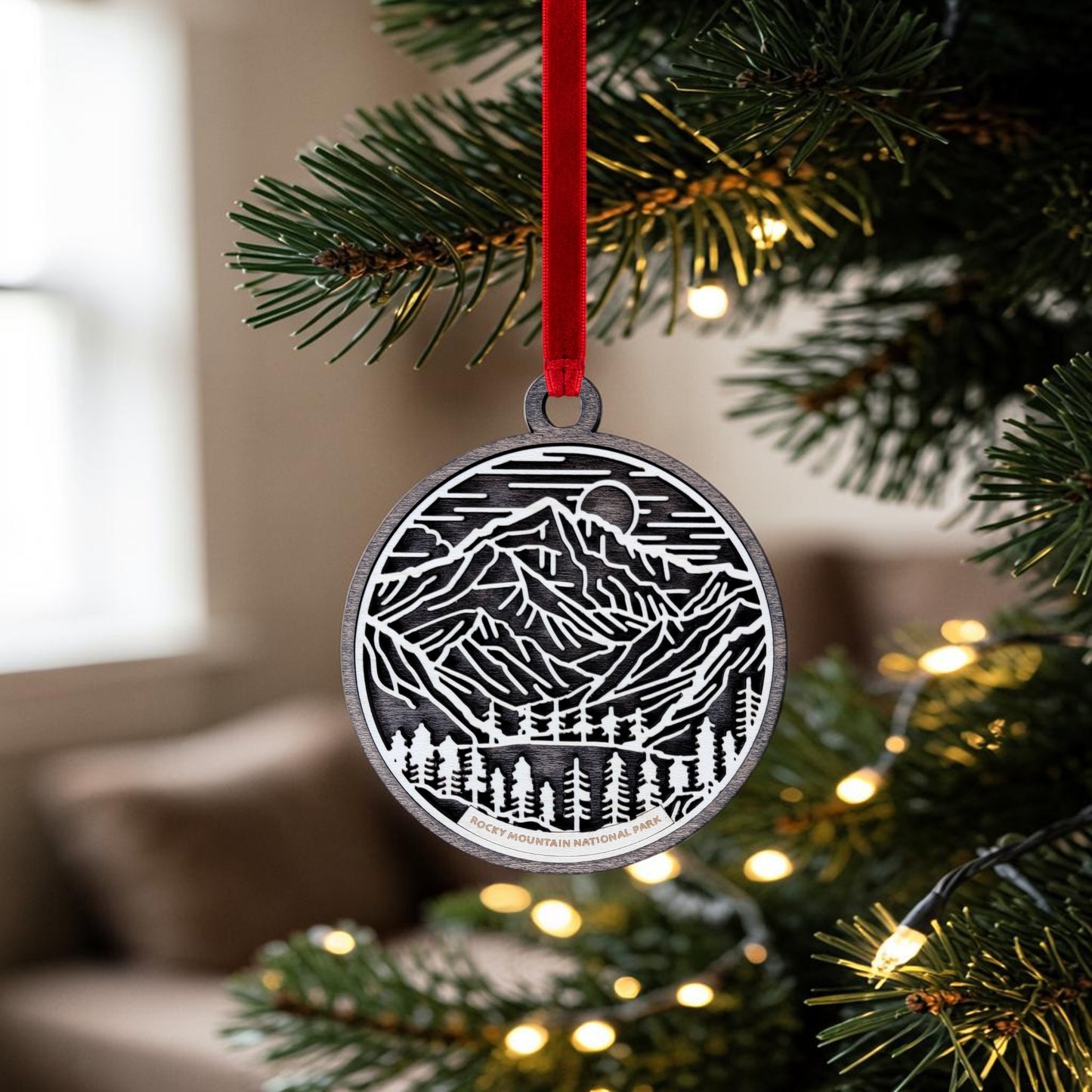 The Mirabilia Boutique Rocky Mountain National Park Line Art Christmas Ornament, featuring a metallic mountain and forest design, hangs from a red ribbon on a Christmas tree adorned with white lights.