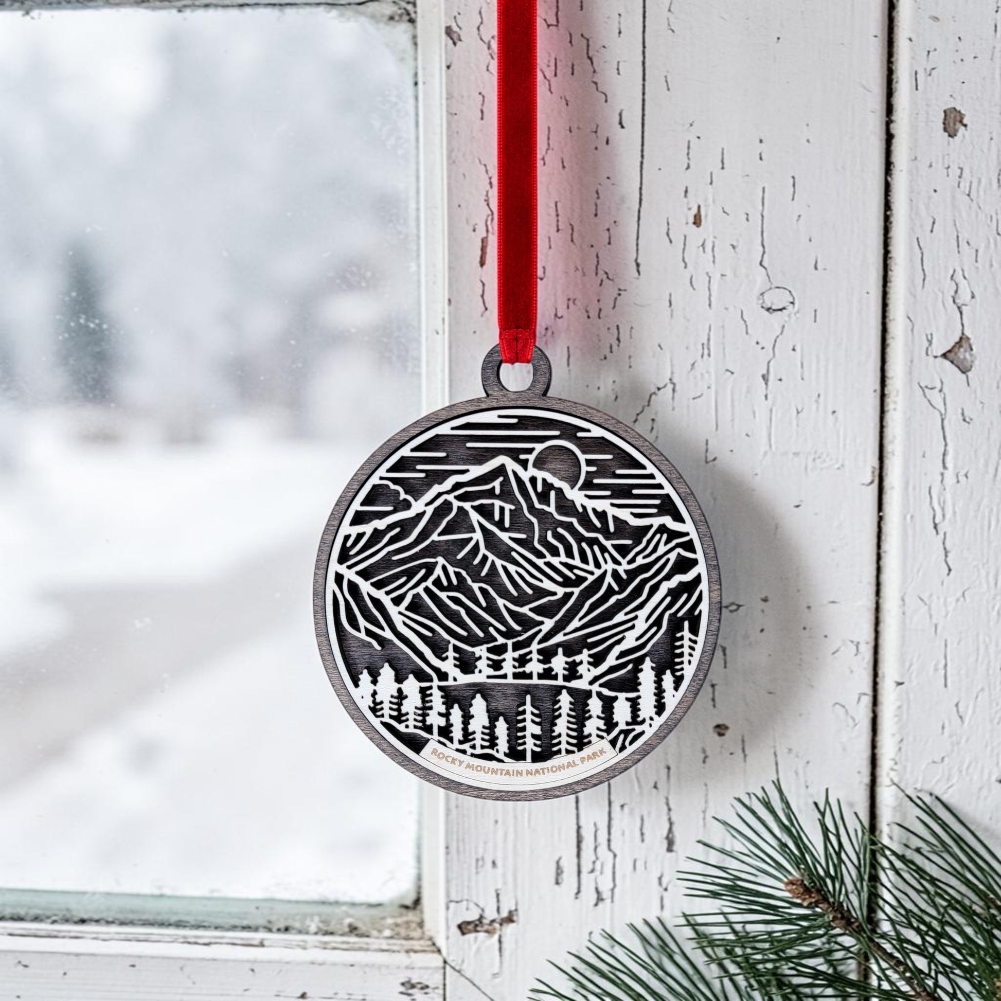 The Rocky Mountain National Park Line Art Christmas Ornament by Mirabilia Boutique, a round wooden piece featuring an intricate mountain and forest design, hangs from a red ribbon on a rustic white window. Outside, snow and pine branches evoke a wintry scene inspired by the iconic national park.