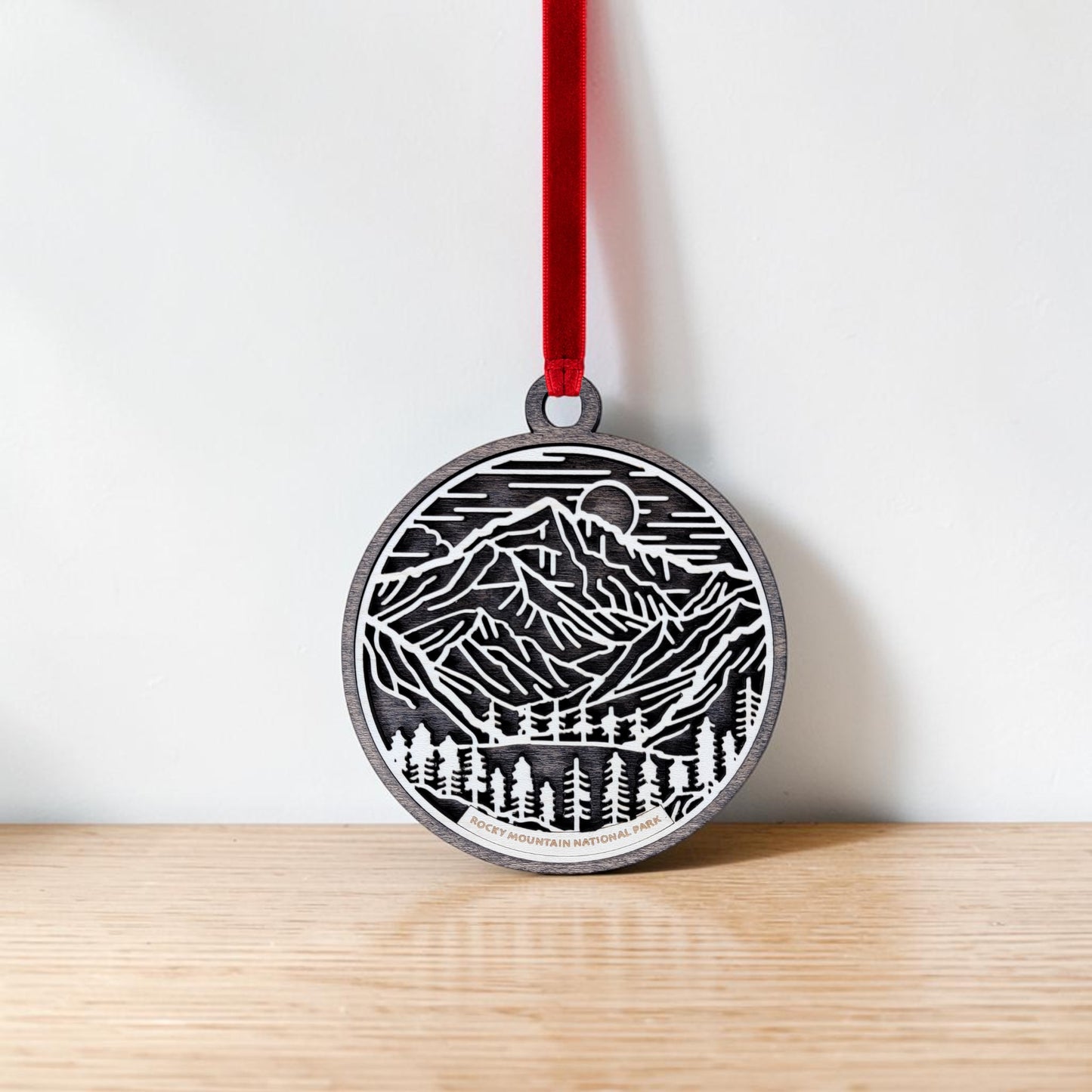 The Rocky Mountain National Park Line Art Christmas Ornament by Mirabilia Boutique—a round wooden decoration adorned with delicate mountain and forest line art—hangs from a red ribbon against a plain white wall and light wooden surface, capturing the tranquil beauty of the park.