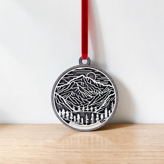 The Rocky Mountain National Park Line Art Christmas Ornament by Mirabilia Boutique—a round wooden decoration adorned with delicate mountain and forest line art—hangs from a red ribbon against a plain white wall and light wooden surface, capturing the tranquil beauty of the park.