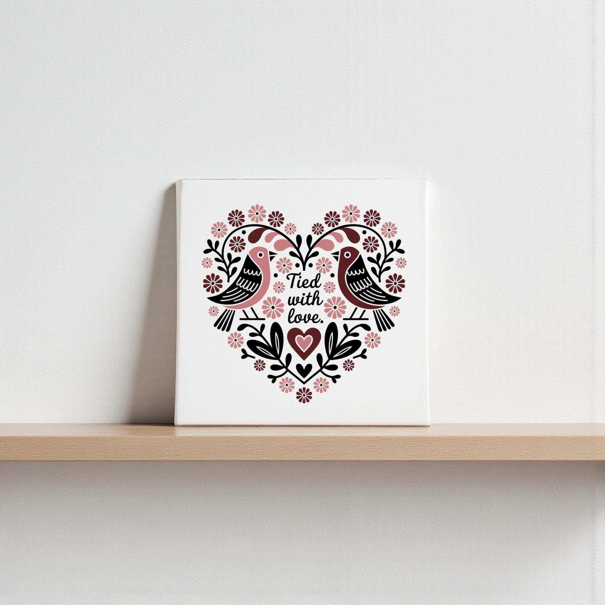 The Tied With Love Folk Art Valentine Canvas by Mirabilia Boutique displays two lovebirds in a heart of Scandinavian floral motifs and the phrase “Tied with love”—perfect for adding unique romance to your wall décor.