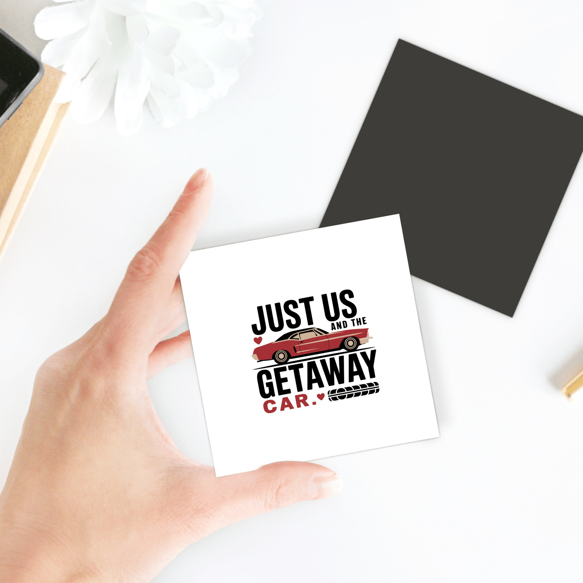 A hand holds a Romantic Getaway Car Magnet by Mirabilia Boutique, featuring a red retro car and the phrase "JUST US AND THE GETAWAY CAR." A flower, black square, and notebook are seen on a white surface in the background.