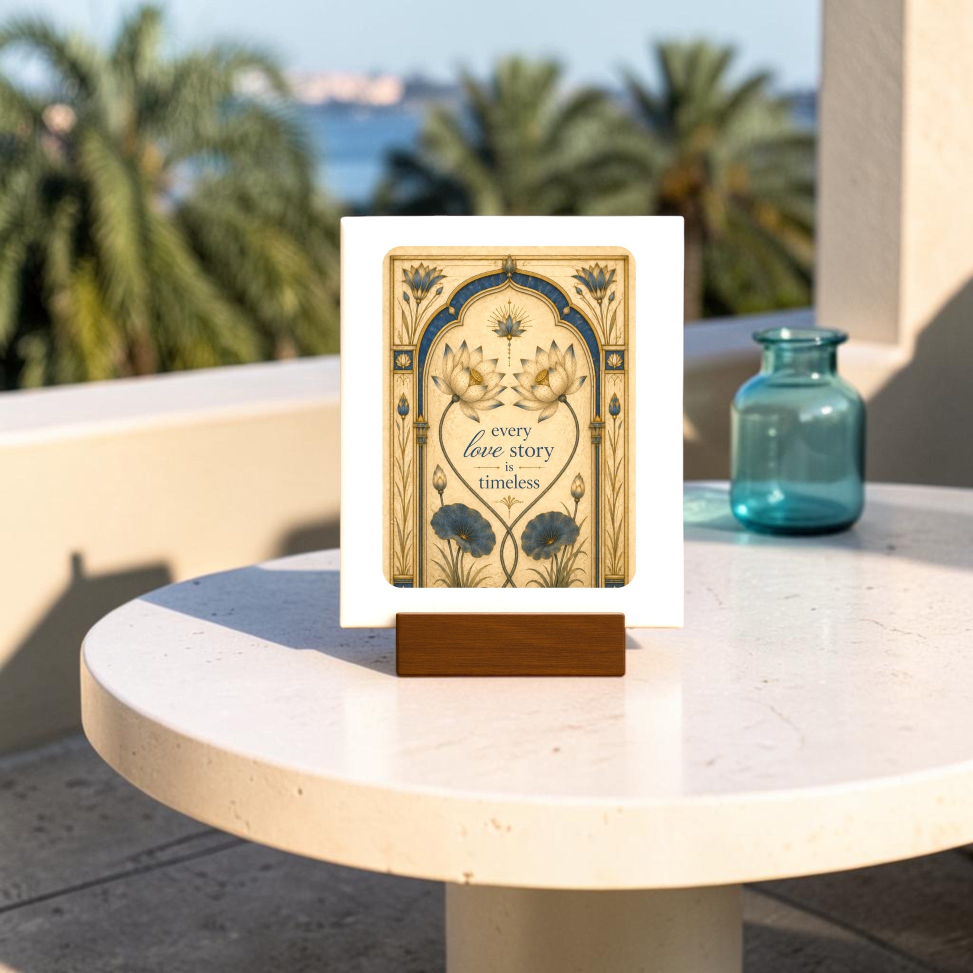 The Mirabilia Boutique “Every Love Story Is Timeless | Lotus Mini Sign,” featuring delicate floral artwork and the heartfelt phrase “every love story is timeless,” rests on a round white table outdoors, set against a backdrop of palm trees and shimmering water.