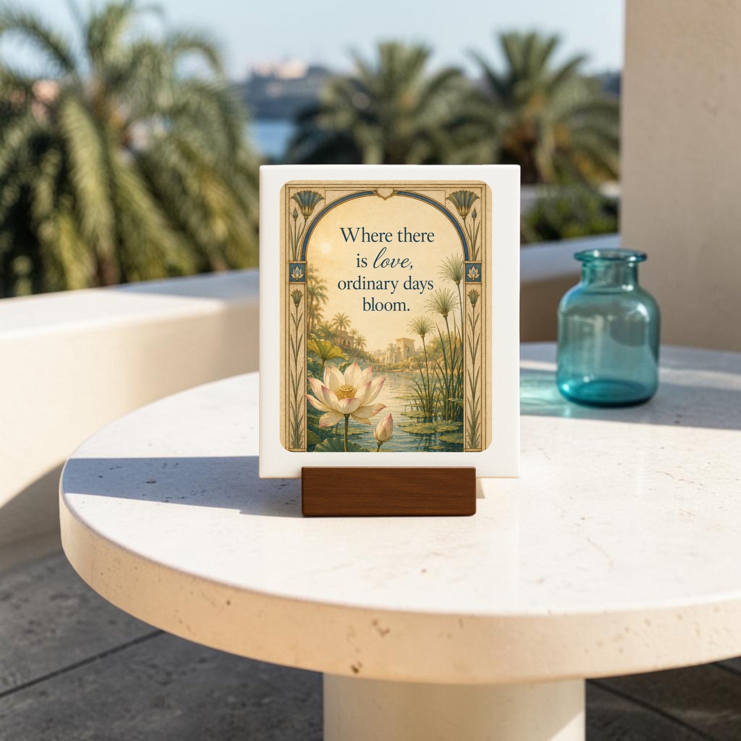 A Where There Is Love | Lotus Mini Sign from Mirabilia Boutique is displayed on a stand atop a round white table with a blue glass vase. The sign features the message, “Where there is love, ordinary days bloom,” and serves as an elegant decorative piece with palm trees and buildings visible in the sunlit background.