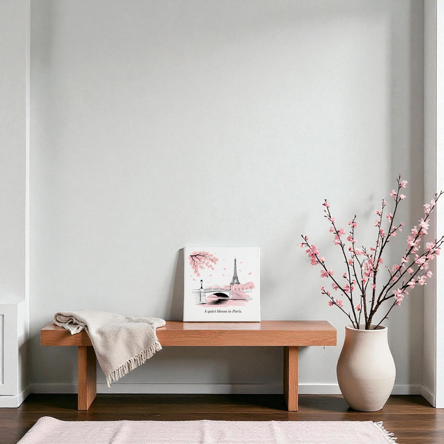 A minimalist room features a wooden bench with a beige throw, Mirabilia Boutique’s Quiet Bloom in Paris Canvas – Cherry Blossom Spring Art, and a large white vase with pink branches, set against a light gray wall and light pink rug.