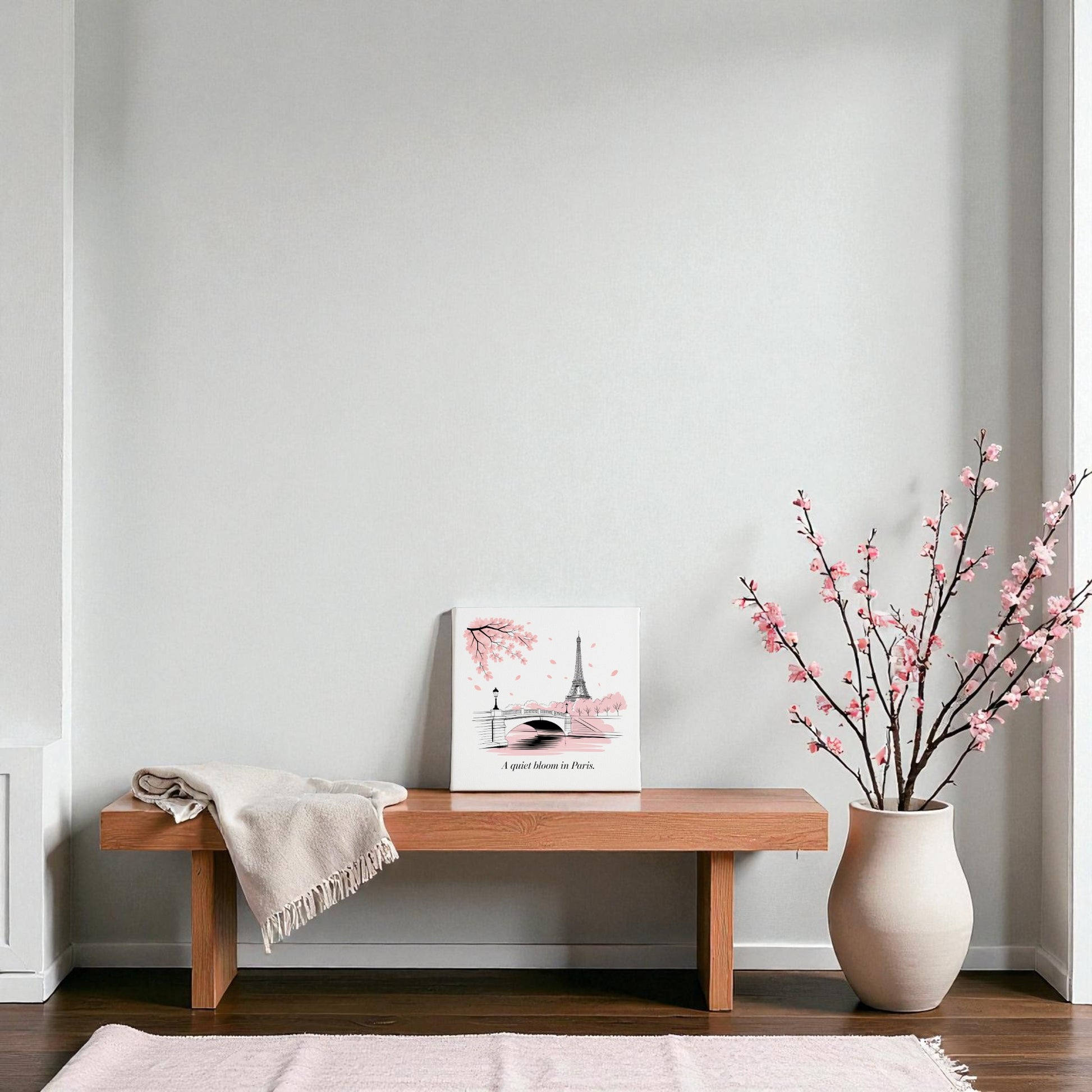 A minimalist room features a wooden bench with a beige throw, Mirabilia Boutique’s Quiet Bloom in Paris Canvas – Cherry Blossom Spring Art, and a large white vase with pink branches, set against a light gray wall and light pink rug.