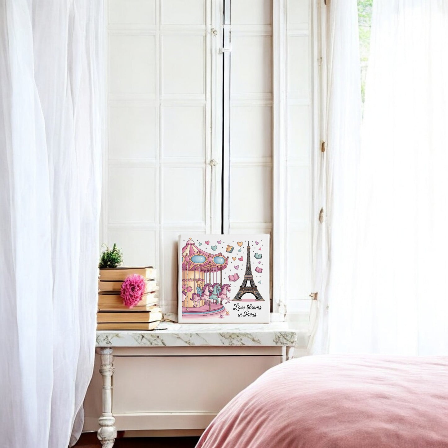 A white room with sheer curtains, a marble-topped table with books and a pink flower, romantic pastel decor, and Mirabilia Boutique’s "Love Blooms in Paris Canvas – Eiffel Tower & Carousel Art." A pink bedspread appears in the foreground.