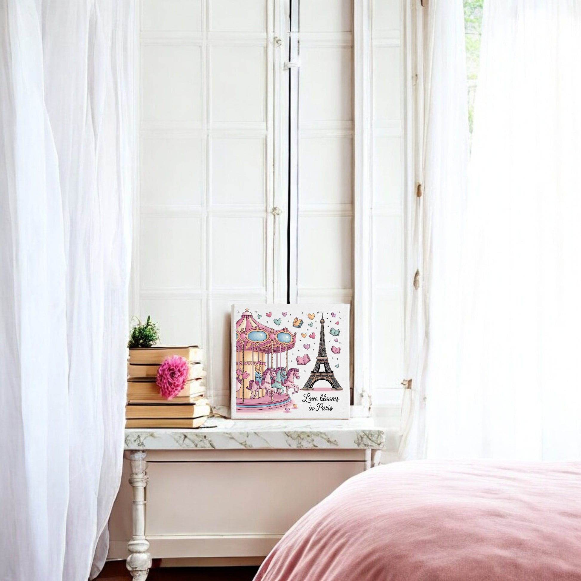 A white room with sheer curtains, a marble-topped table with books and a pink flower, romantic pastel decor, and Mirabilia Boutique’s "Love Blooms in Paris Canvas – Eiffel Tower & Carousel Art." A pink bedspread appears in the foreground.