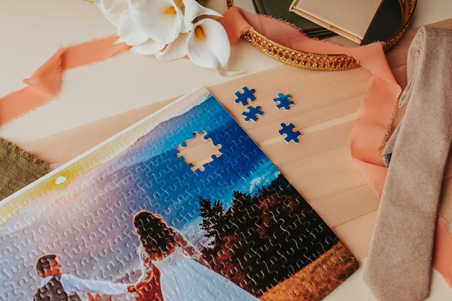 A nearly finished Mirabilia Boutique Custom Wedding Jigsaw Puzzle | 300-Piece Photo Keepsake, featuring a couple outdoors with one piece missing, is displayed among flowers, ribbons, a gold mirror, and beige cloth on a table. Four loose pieces sit nearby.