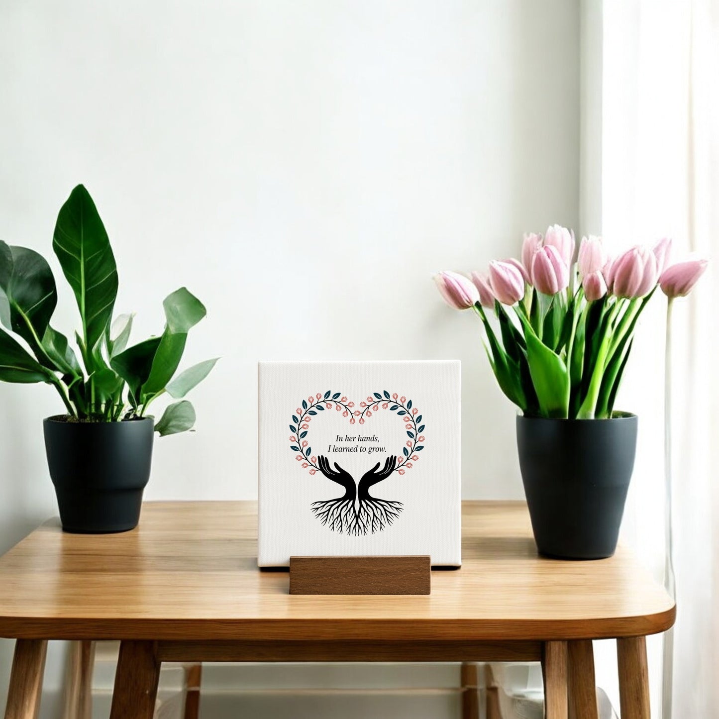 A small wooden table displays two potted plants and Mirabilia Boutique’s “In Her Hands Mini Sign”—a thoughtful Mother’s Day gift featuring a tree design and the words, “In her hands, I learned to grow,” against a light wall and sheer curtain.