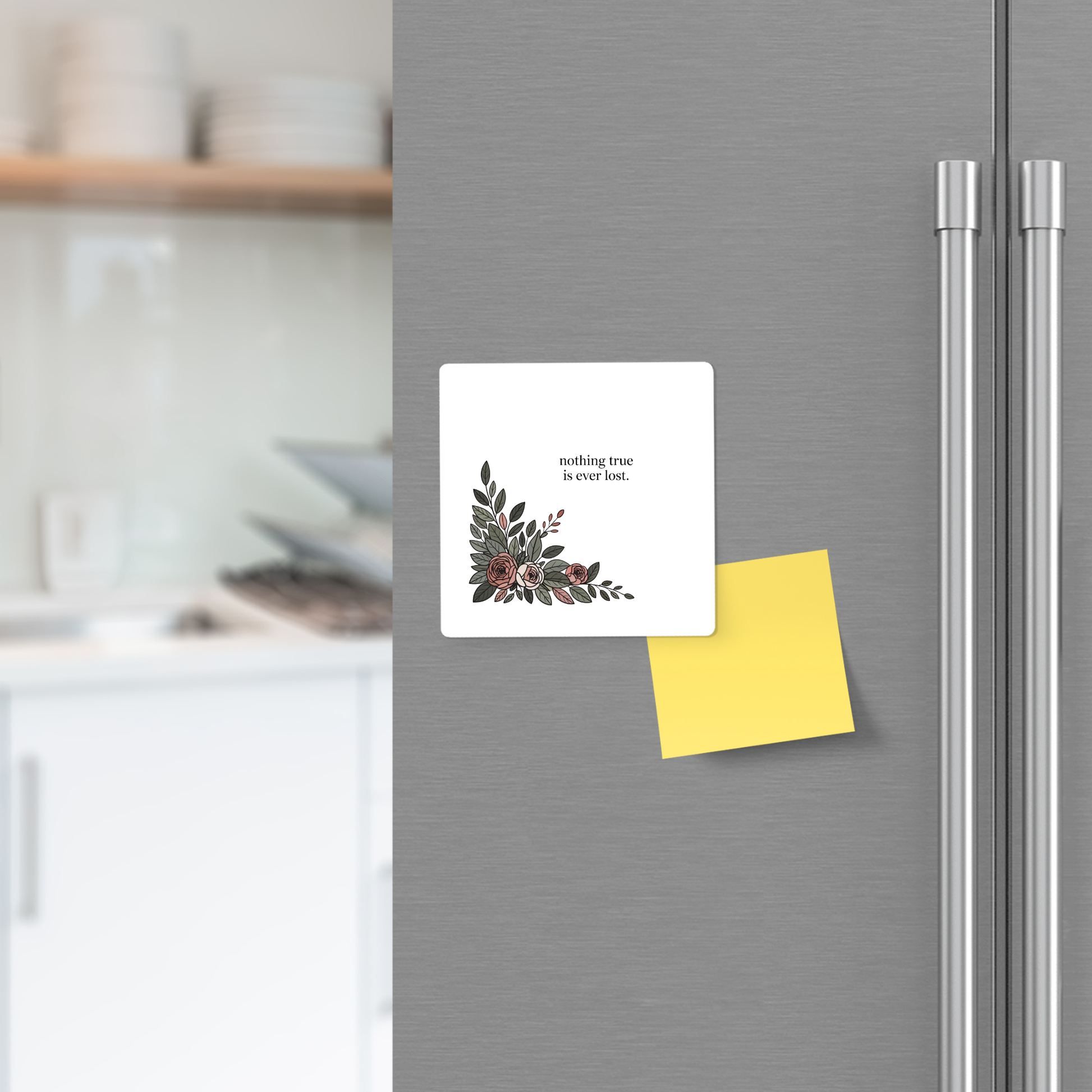 The Mirabilia Boutique "Nothing True Is Ever Lost Floral Magnet" holds a card with its uplifting message and a yellow sticky note on a refrigerator door, while blurred kitchen shelves and dishes appear in the background.