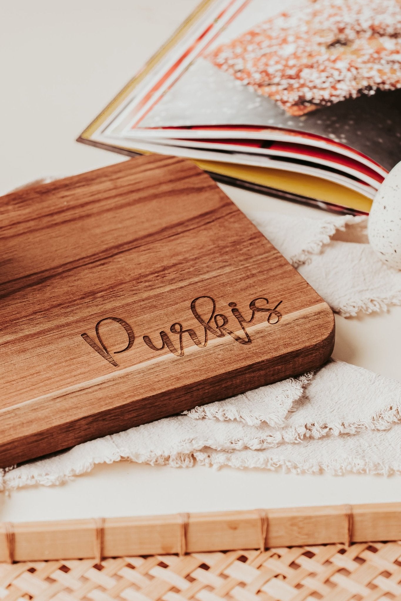 The Mirabilia Boutique Engraved Acacia Cutting Board (14x8) rests on a white cloth with an open cookbook and ceramic cup—an ideal personalized kitchen gift.