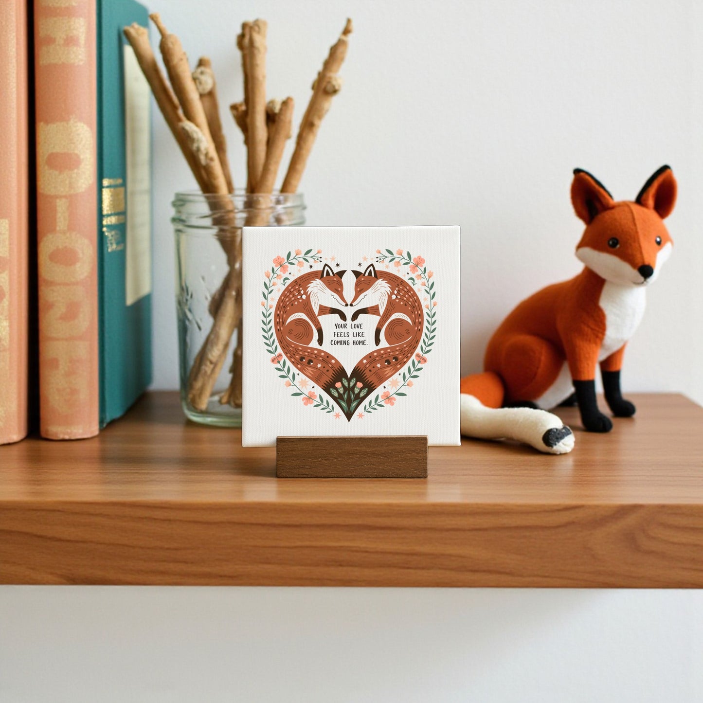 A small wooden shelf displays books, a glass jar with sticks, a felt fox figurine, and the Fox Love Mini Canvas by Mirabilia Boutique—a “Your Love Feels Like Home” art piece perfect for cottagecore woodland or rustic romance decor.