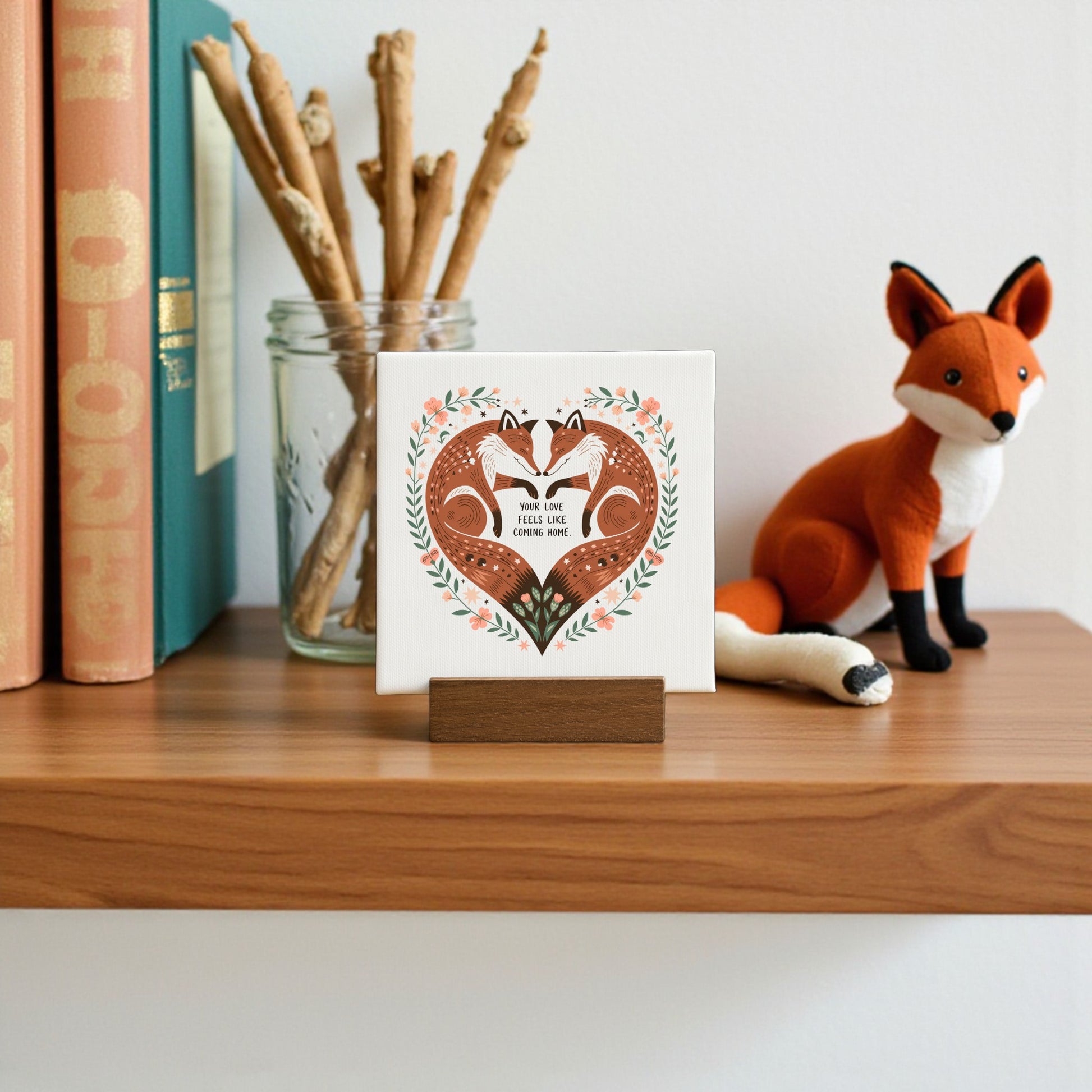 A small wooden shelf displays books, a glass jar with sticks, a felt fox figurine, and the Fox Love Mini Canvas by Mirabilia Boutique—a “Your Love Feels Like Home” art piece perfect for cottagecore woodland or rustic romance decor.