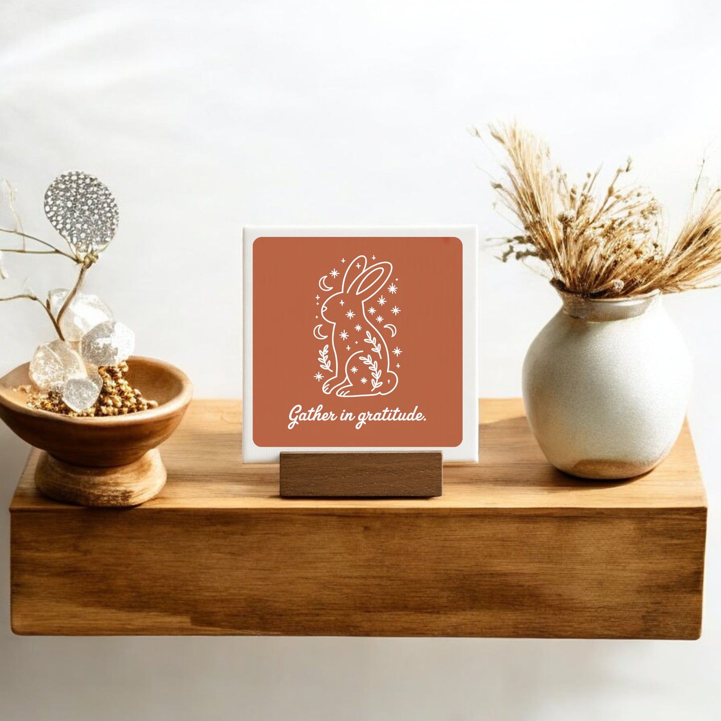 A wooden shelf displays the Mirabilia Boutique "Gather in Gratitude Mini Canvas Sign – 4x4 Bunny," featuring a white bunny outline, stars, flowers, and "Gather in gratitude." Vases with dried grasses and gold beads complete this cozy decor arrangement.