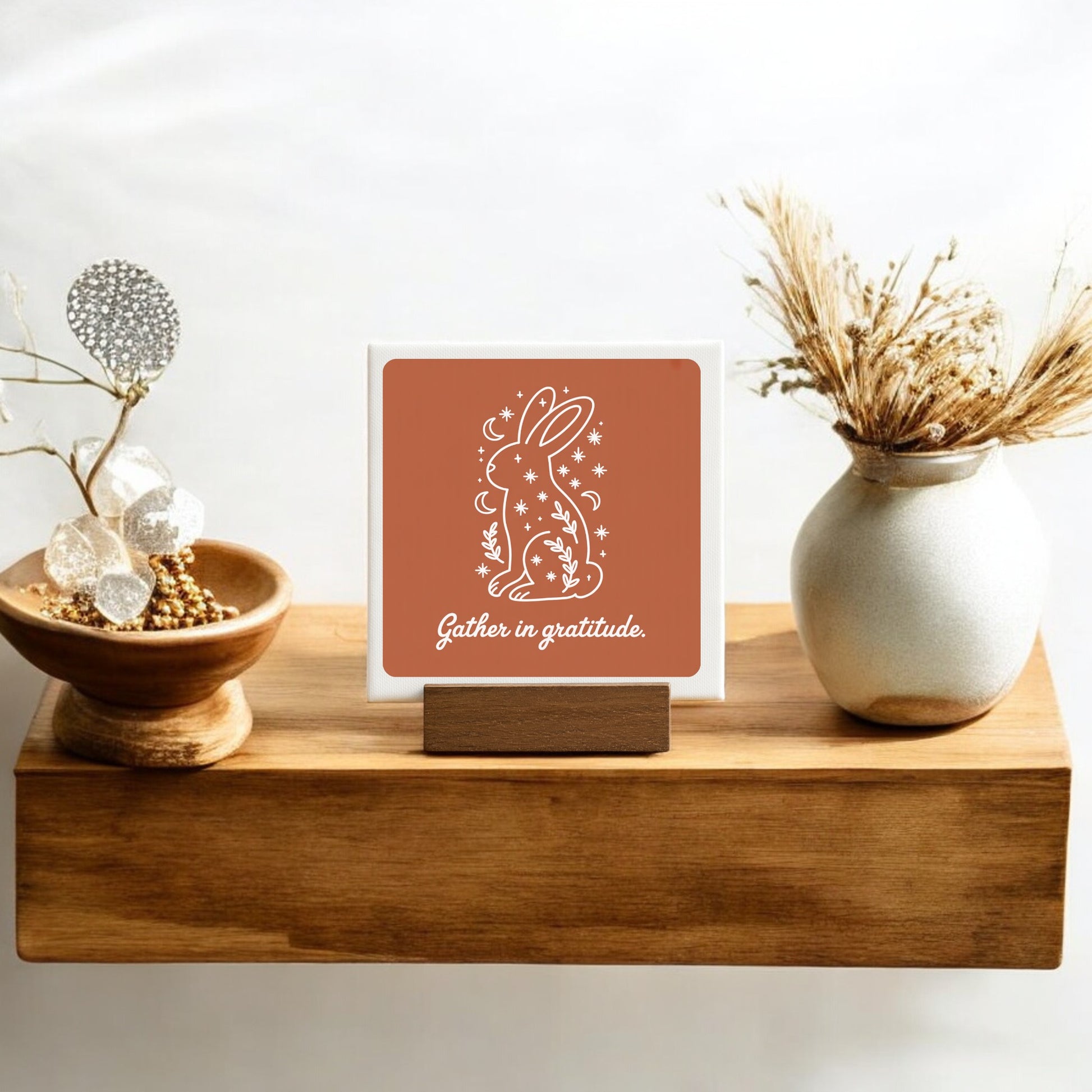 A wooden shelf displays the Mirabilia Boutique "Gather in Gratitude Mini Canvas Sign – 4x4 Bunny," featuring a white bunny outline, stars, flowers, and "Gather in gratitude." Vases with dried grasses and gold beads complete this cozy decor arrangement.