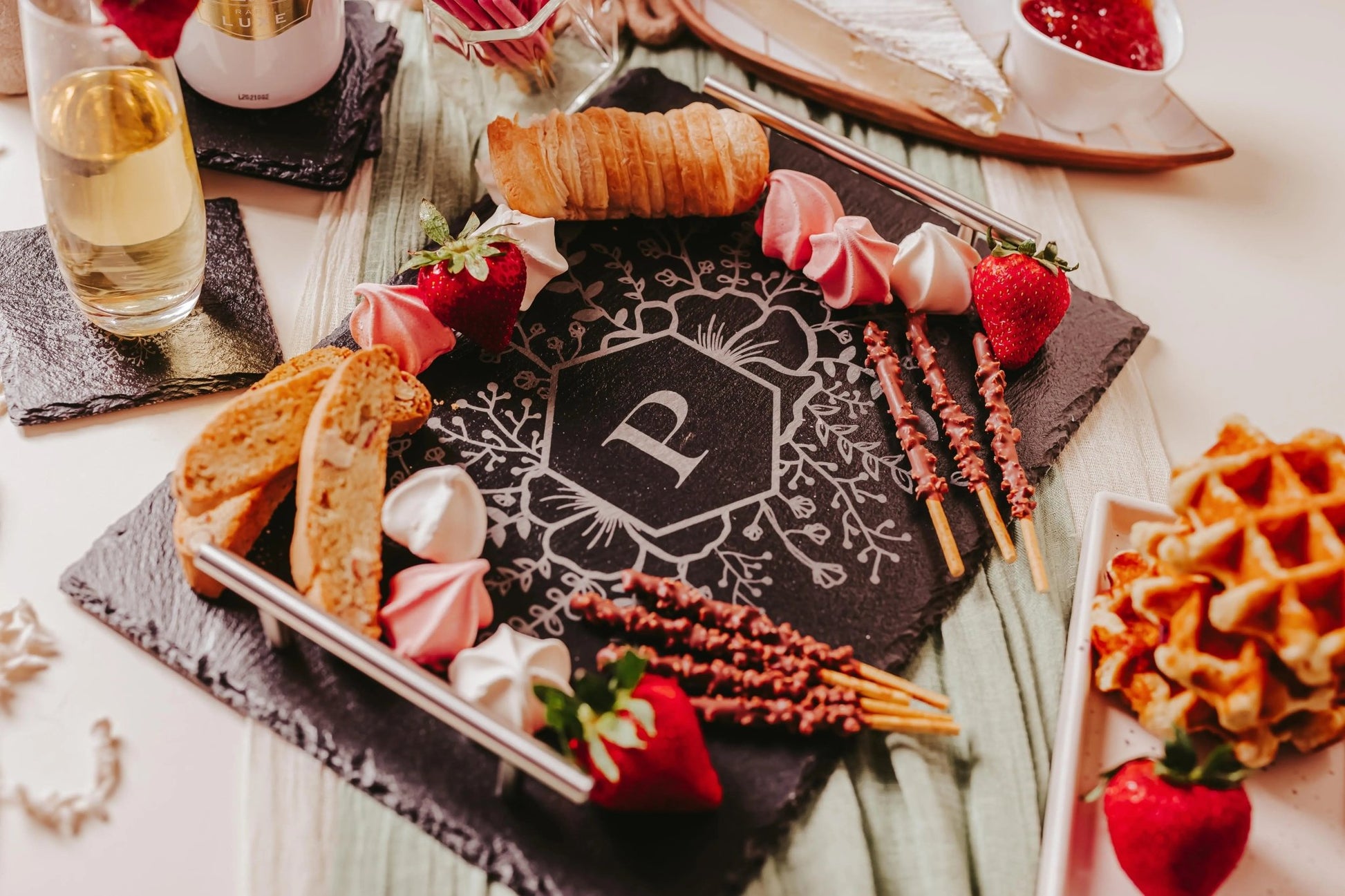 The Mirabilia Boutique Personalized Engraved Slate Serving Tray with Handles features a floral “P” monogram and is elegantly styled with strawberries, meringues, biscotti, chocolate sticks, a croissant, waffles, and white wine—a perfect custom gift.