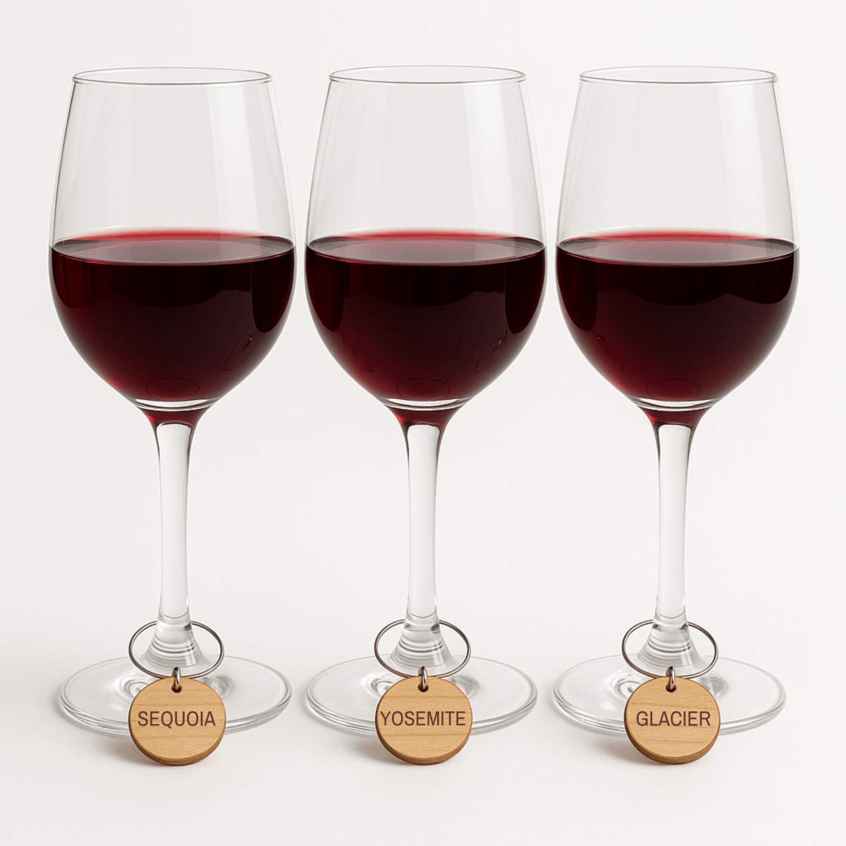 Three glasses of red wine feature Mirabilia Boutique’s Mountain Majesty National Park Wine Charms—round wooden tags labeled "SEQUOIA," "YOSEMITE," and "GLACIER"—adding national park flair to your gathering.