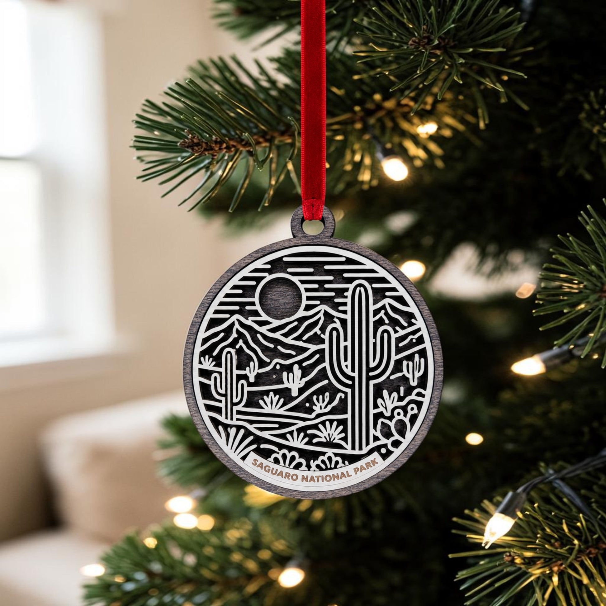 The Saguaro National Park Line Art Christmas Ornament by Mirabilia Boutique features a wooden desert scene in delicate line art, hanging from a festive red ribbon on a sparkling, decorated Christmas tree—perfect as a distinctive desert landscape keepsake for the holiday season.