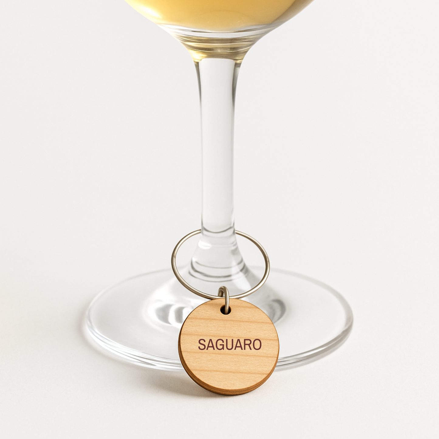 A clear wine glass filled with white wine is adorned with a Desert Dreaming Wine Charm from Mirabilia Boutique, featuring a wooden "SAGUARO" label, shown against a plain white background.