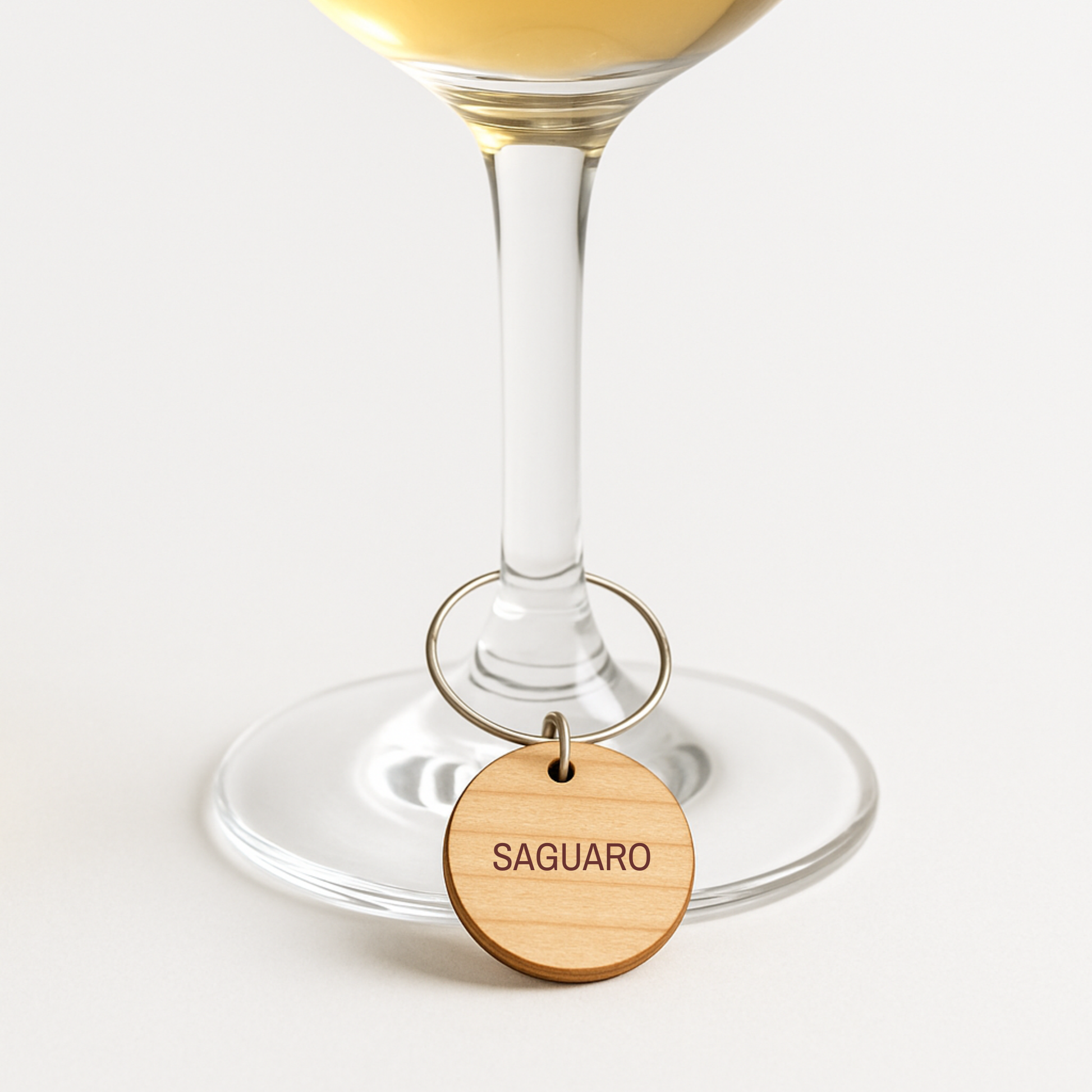 A clear wine glass filled with white wine is adorned with a Desert Dreaming Wine Charm from Mirabilia Boutique, featuring a wooden "SAGUARO" label, shown against a plain white background.