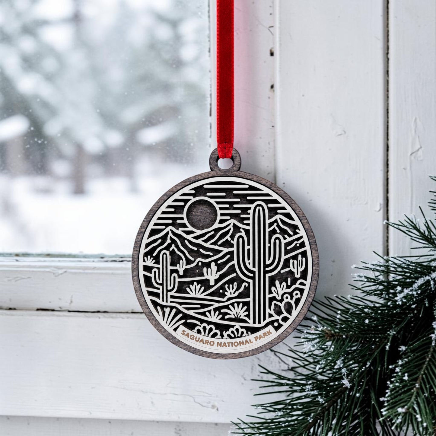 A Saguaro National Park Line Art Christmas Ornament by Mirabilia Boutique, featuring a round wooden design with a desert scene of cacti and mountains, hangs from a red ribbon on a white window. Pine branches are nearby, and snow can be seen outside. The ornament bears the words "Saguaro National Park.