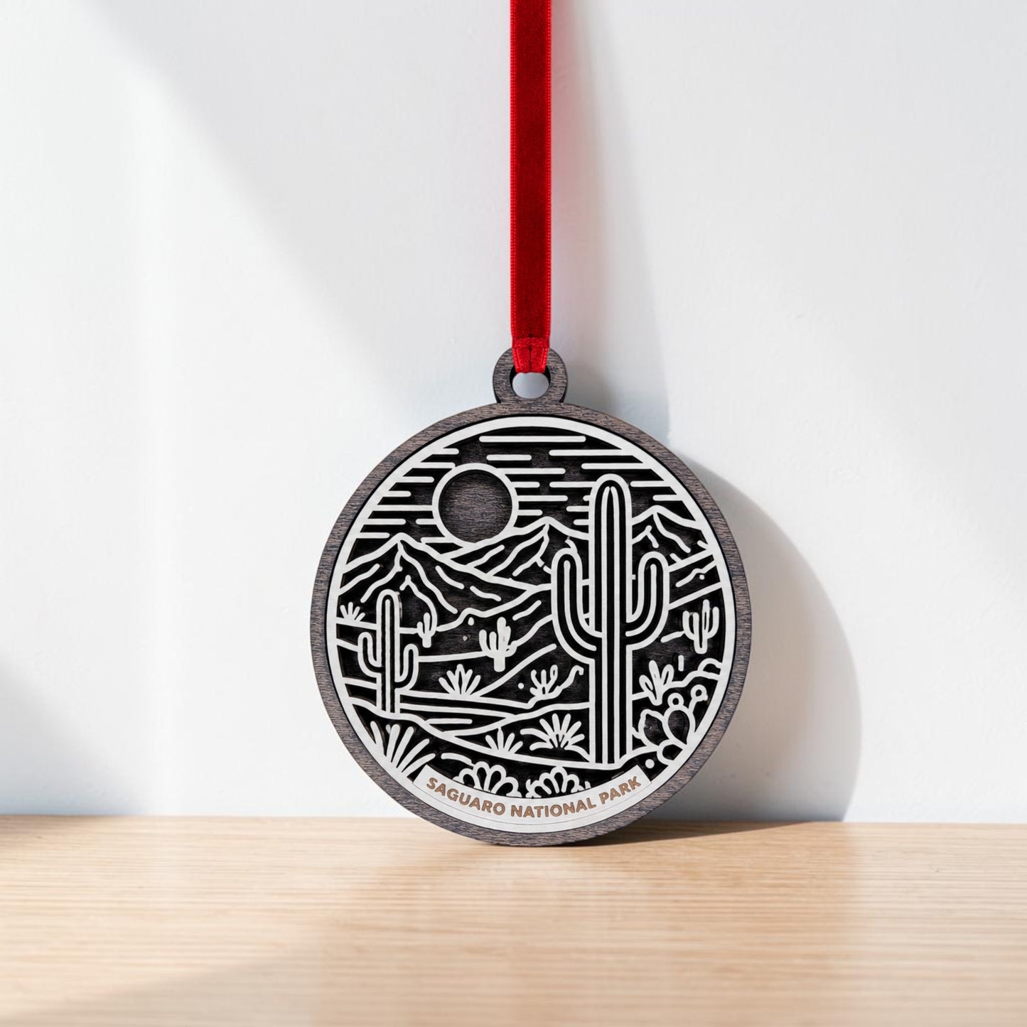 The Saguaro National Park Line Art Christmas Ornament by Mirabilia Boutique features a round design with a red ribbon and an engraved desert landscape of cacti, mountains, and a sun or moon. This ornament is a stunning keepsake for desert lovers, with “Saguaro National Park” inscribed at the base, and is pictured resting against a white wall on a wooden surface.