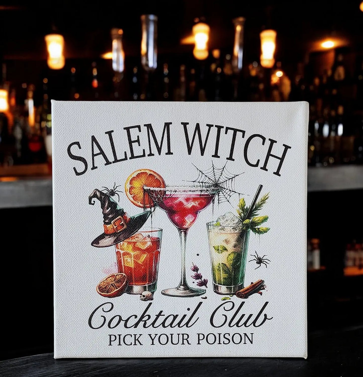 A canvas titled "Salem Witch Cocktail Club Sign - Spooky Halloween Bar Decor" from Mirabilia Boutique. The design features illustrated witchy cocktails adorned with witch hats, spider webs, and citrus slices, set against a dimly lit bar background filled with mysterious bottles to perfectly capture the Halloween spirit.