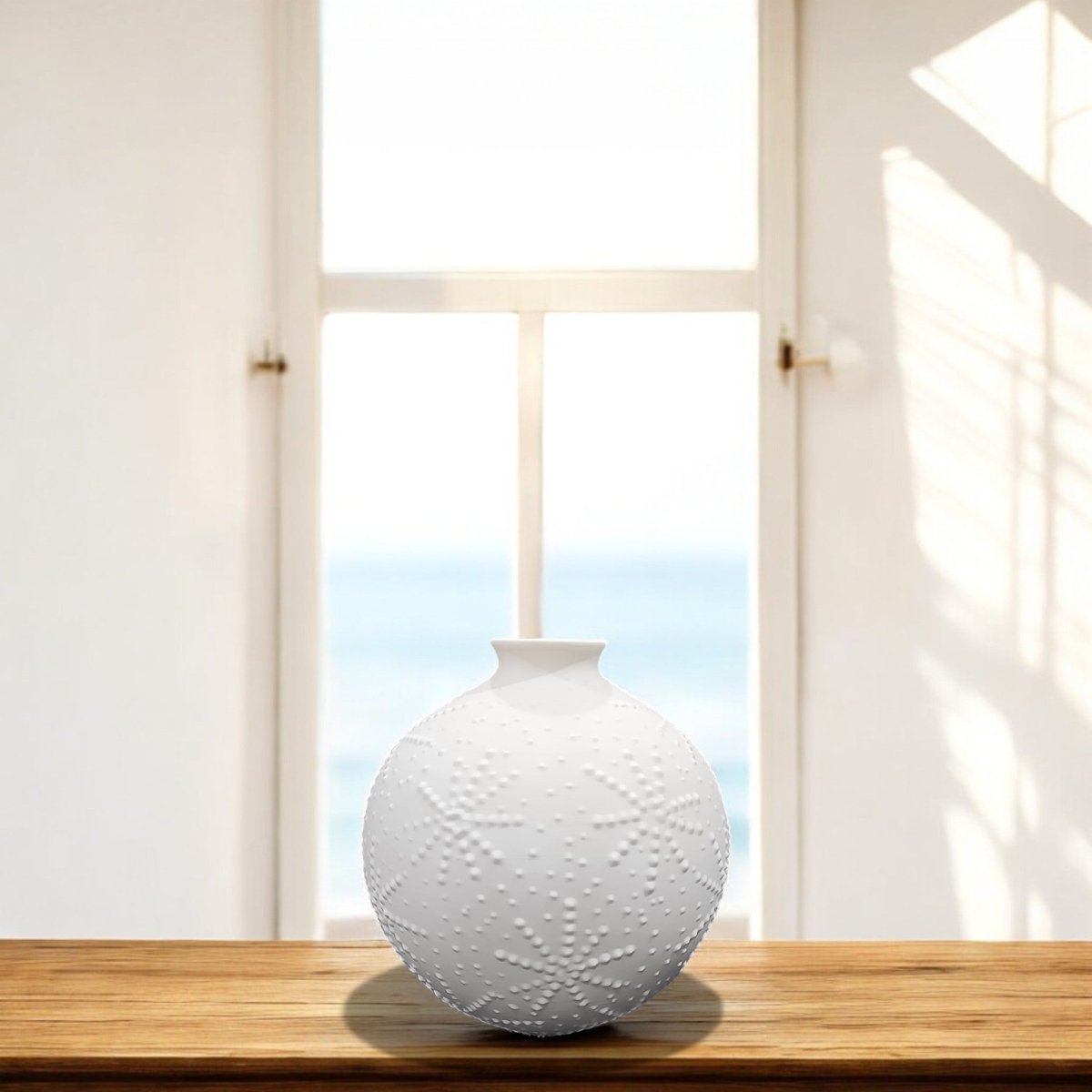 The Mirabilia Boutique Sand Dollar Textured Vase in Matte White sits on a wooden table by a sunlit window, enhancing the coastal decor with an ocean view backdrop.