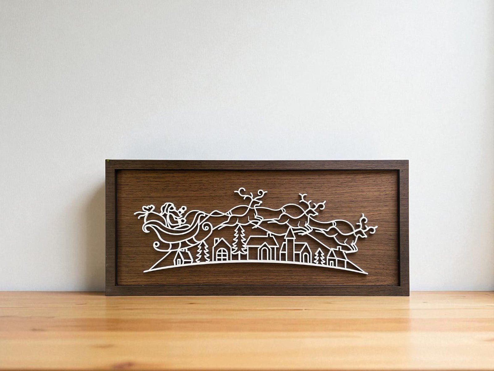 The Santa Sleigh Laser Cut Sign by Mirabilia Boutique features a modern wooden frame with Santa’s sleigh and reindeer over a village, making it perfect minimalist Christmas wall art for your home decor.