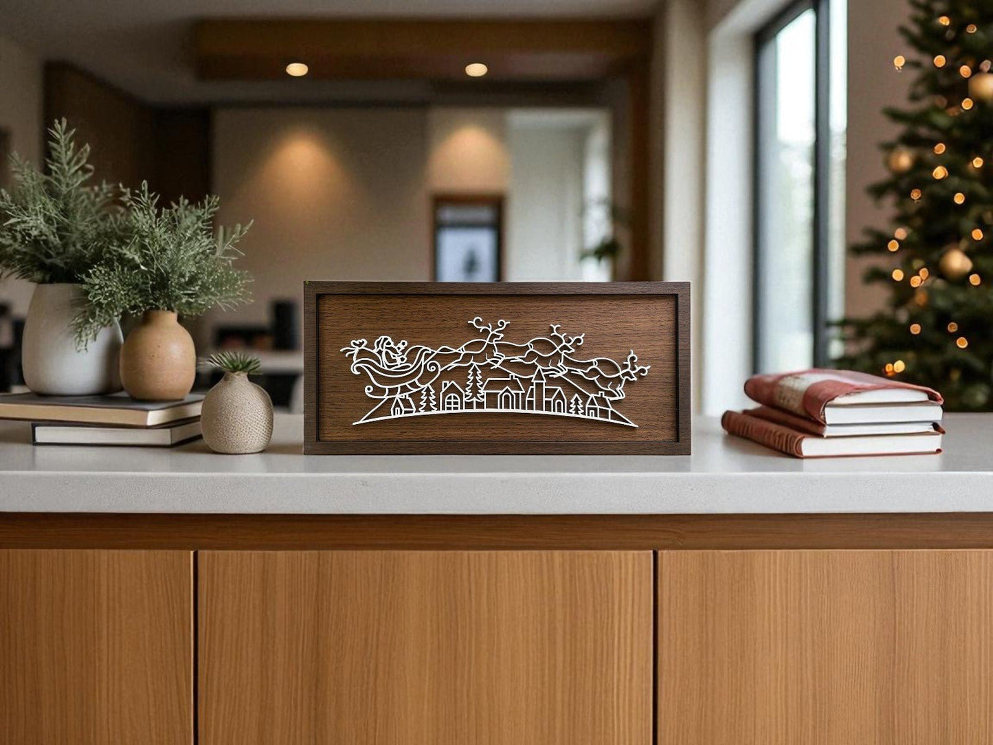 The Santa Sleigh Laser Cut Sign by Mirabilia Boutique adds a modern touch to your kitchen island, styled with a small plant, two vases, and books. Minimalist holiday decor and a Christmas tree complete the festive scene.