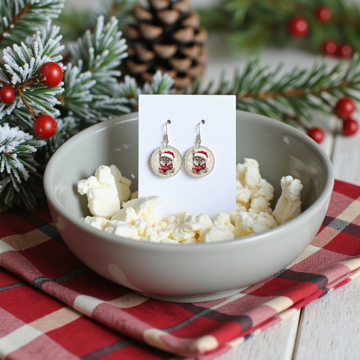 A bowl of popcorn holds a card with Mirabilia Boutique's Santa Wolf Earrings | Woodland Christmas Jewelry Gift, set on red plaid and surrounded by pine branches, berries, and pinecone—perfect hypoallergenic holiday jewelry.