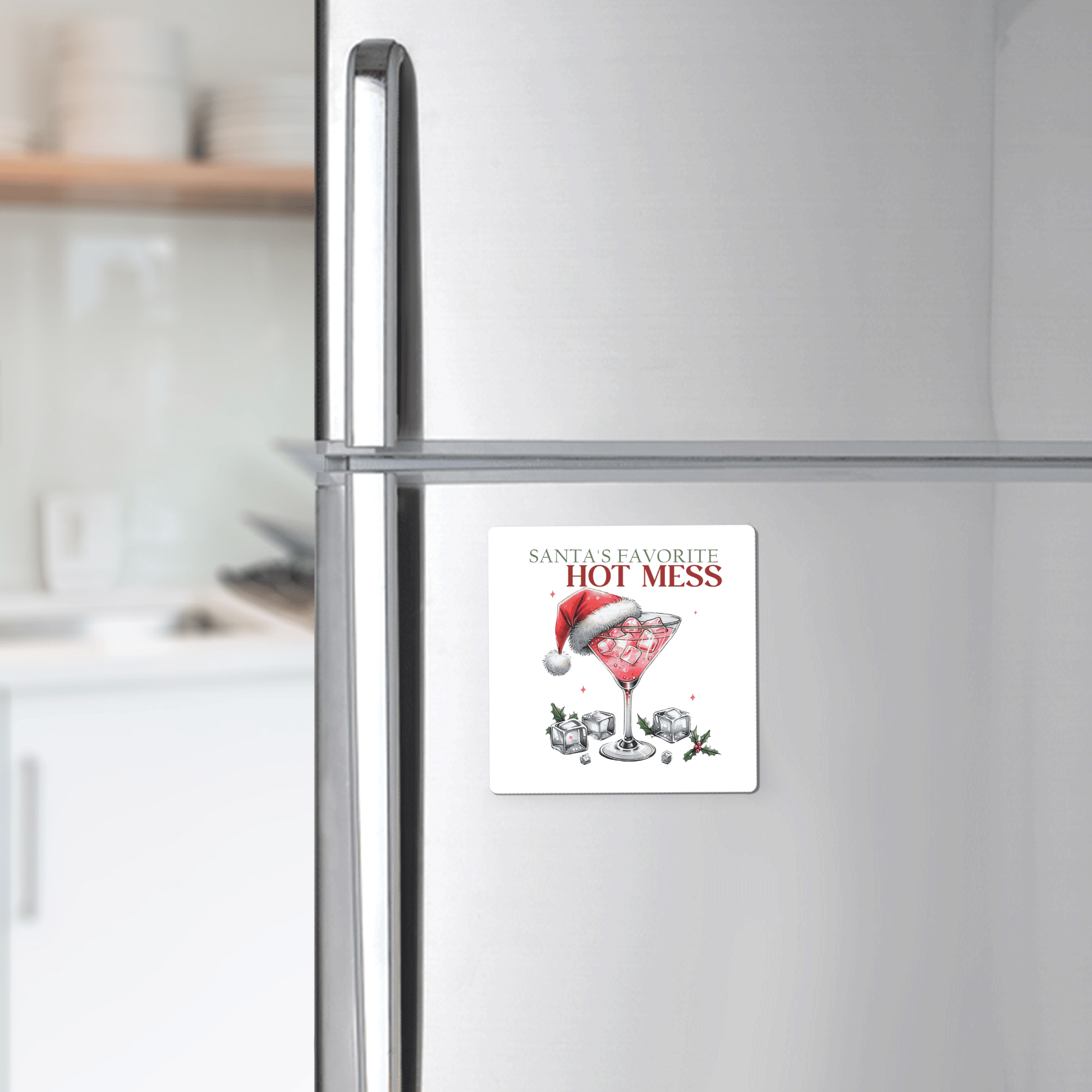 The Santa’s Favorite Hot Mess Magnet by Mirabilia Boutique features "Santa's Favorite Hot Mess" above a cocktail glass with a Santa hat, ice, and holly—a funny holiday fridge decor perfect as a stocking stuffer. Kitchen items blur in the background.