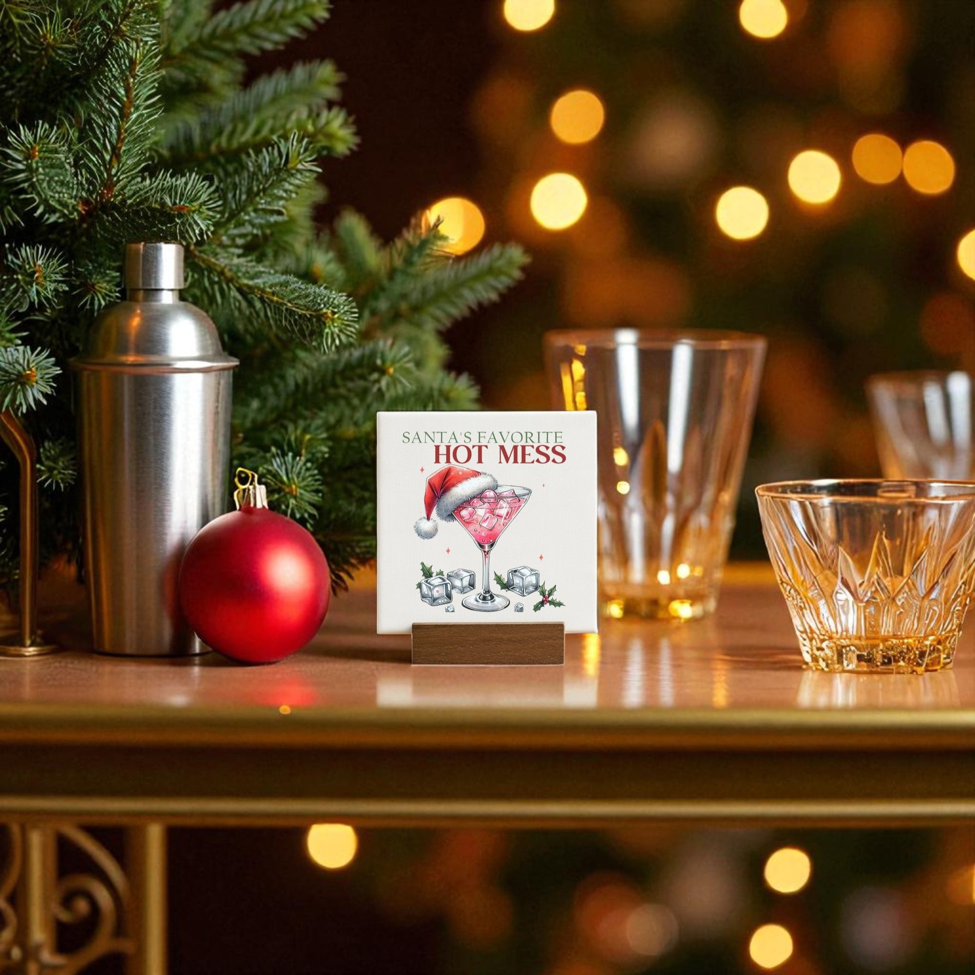 A festive bar scene with Mirabilia Boutique’s “Santa’s Favorite Hot Mess” Mini Sign, holiday glassware, a cocktail shaker, red ornament, tree branch decor, and warm string lights—the perfect funny holiday hostess gift.
