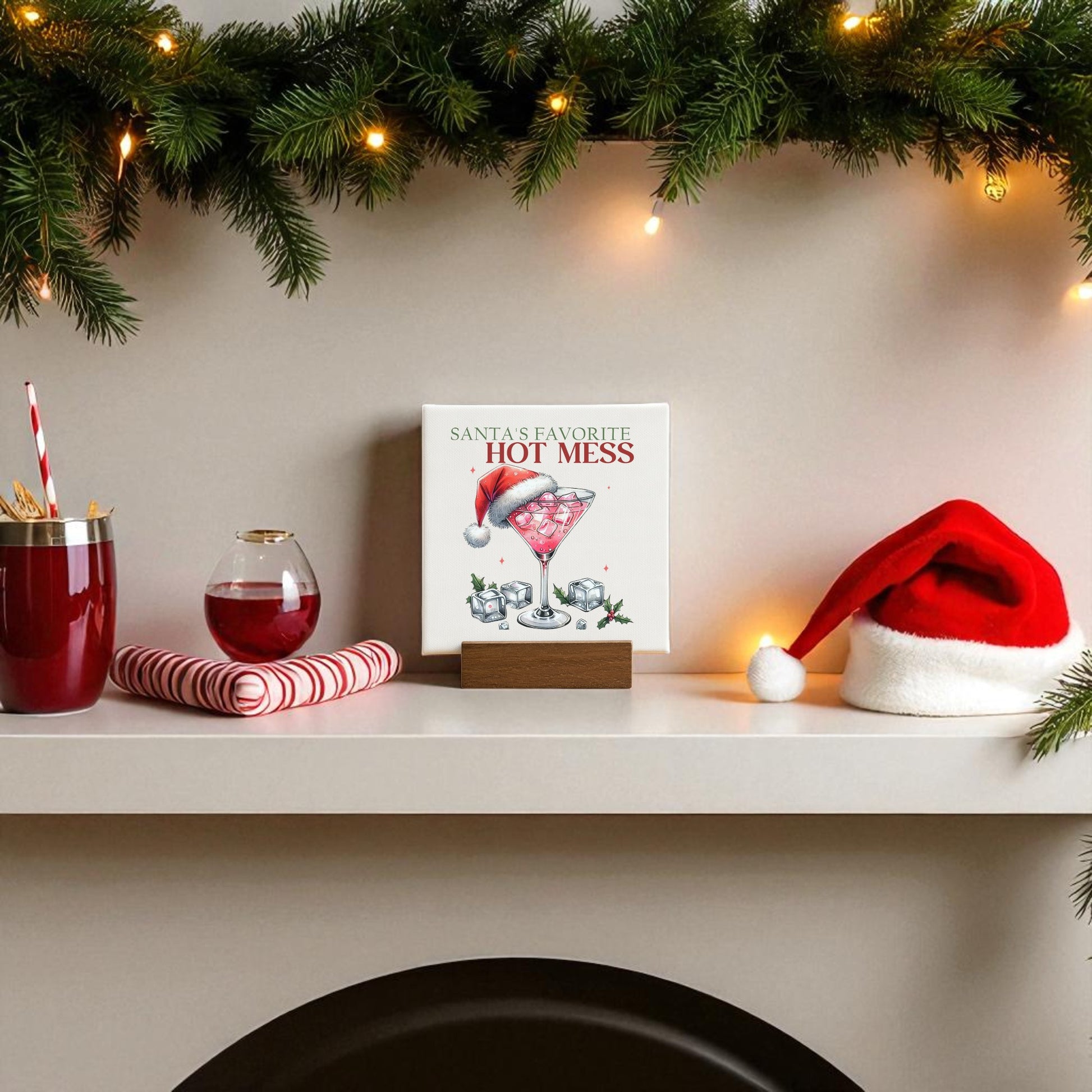 A festive mantel with garland, a Santa hat, red drinks, candy canes, and the "Santa’s Favorite Hot Mess Mini Sign" by Mirabilia Boutique—perfect as quirky Christmas bar decor or a fun holiday hostess gift.