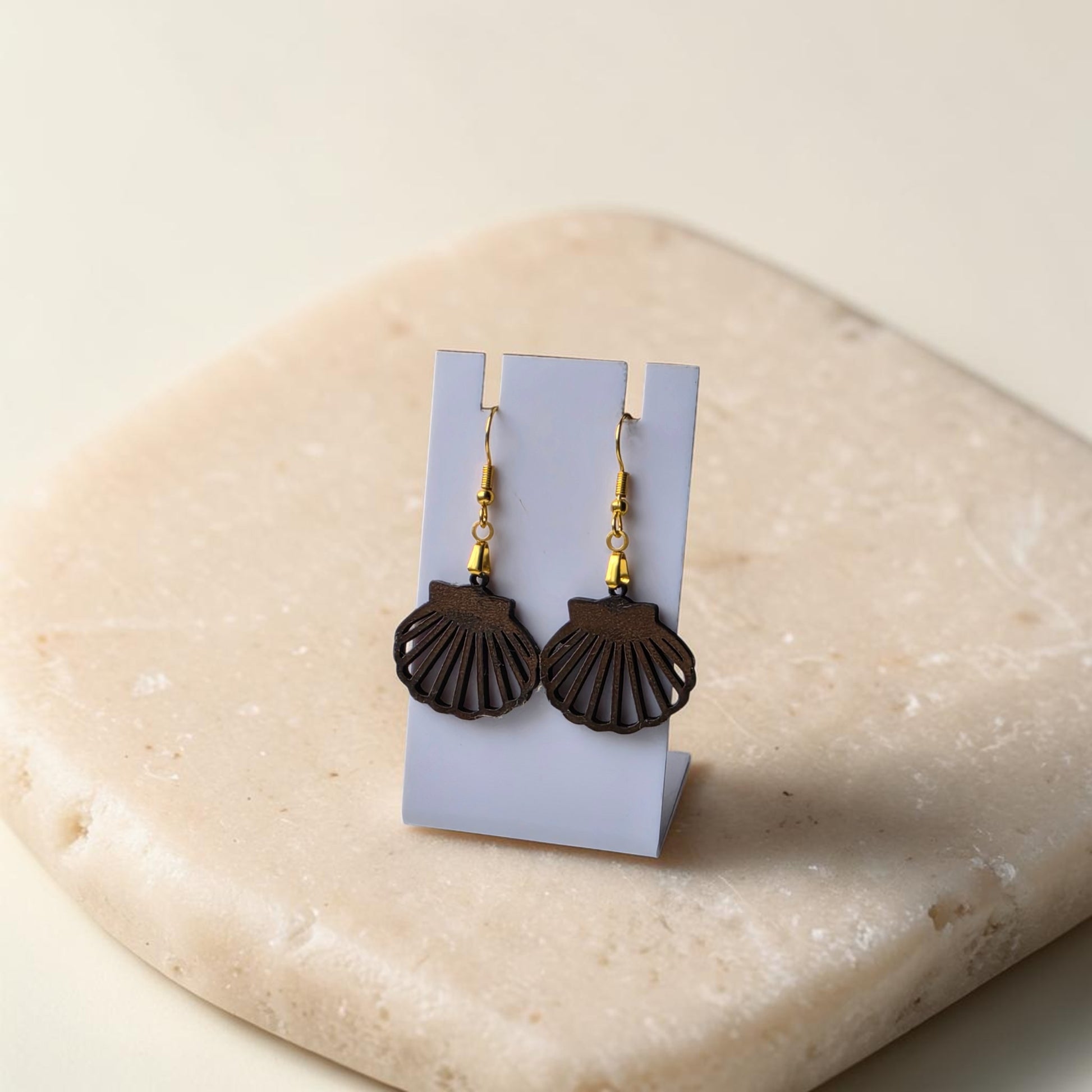 Scallop Shell Earrings | Lasercut Walnut Wood by Mirabilia Boutique feature dark brown walnut shells and gold hooks, displayed on a white card atop a light beige stone—ideal for lovers of coastal jewelry.