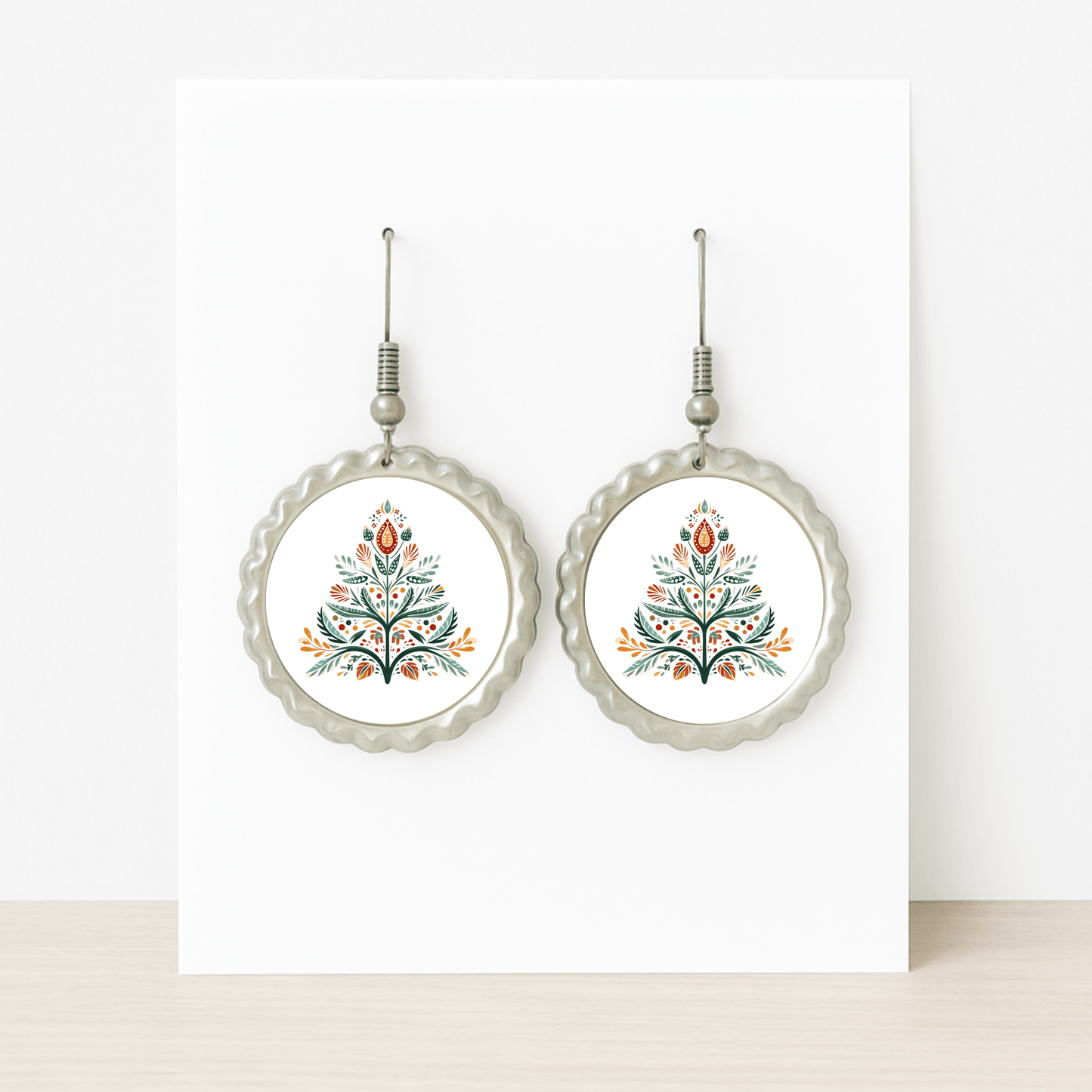 Scandi Christmas Tree Earrings by Mirabilia Boutique: round Nordic-inspired design with scalloped silver edges and a vibrant red, green, yellow, and black floral folk art motif on white.